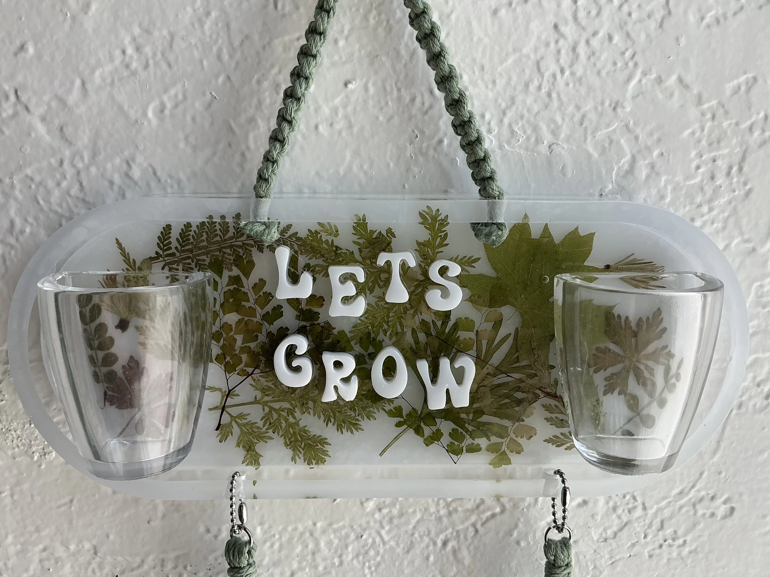 Close front view of the clear rounded resin panel with embedded pressed greenery, raised white “LET’S GROW” letters, and two empty transparent air plant holders.