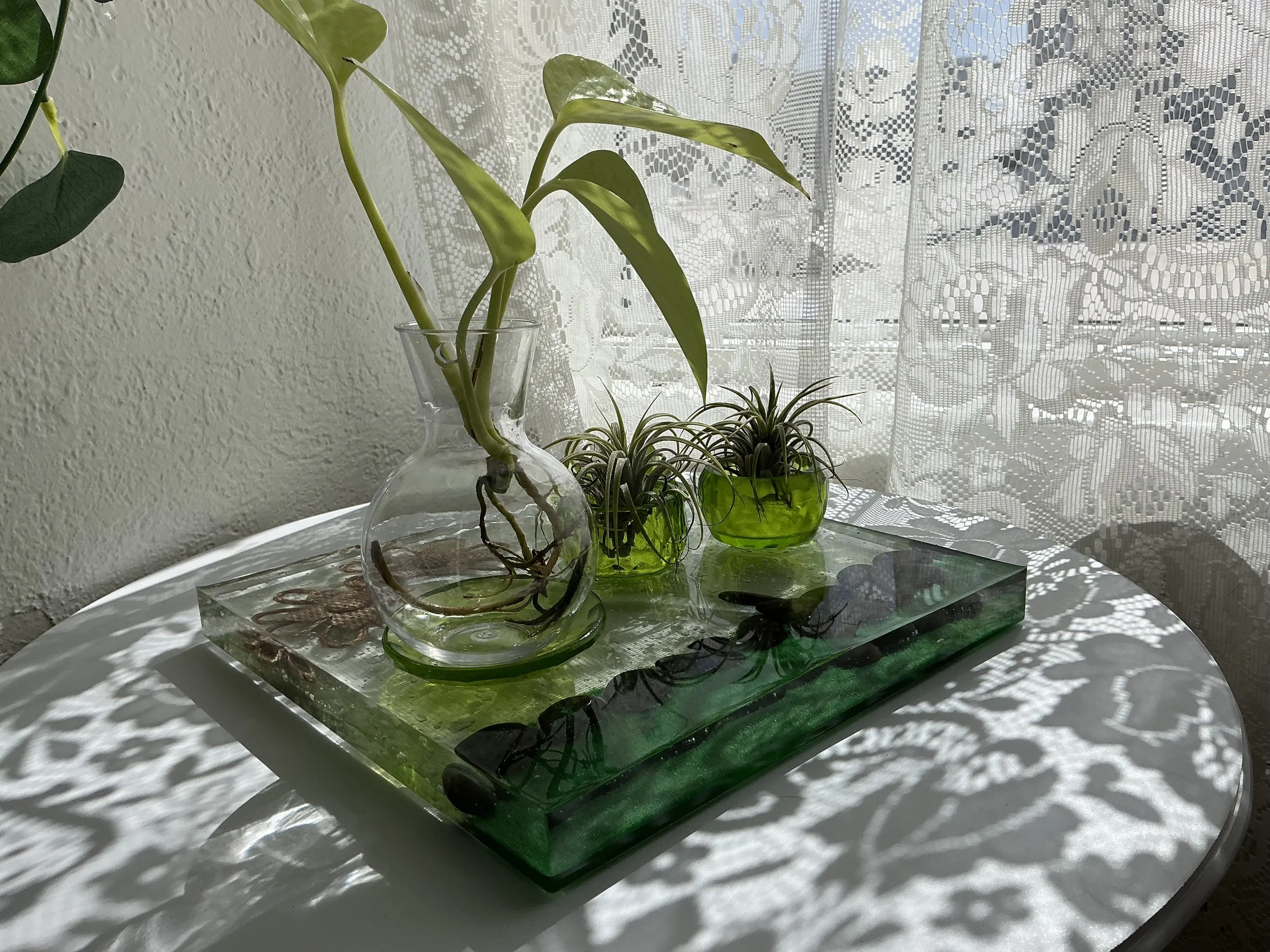 Angled view of the clear resin propagation station with a glass vase, two green air plant holders, an emerald edge lined with black stones, burlap flowers and a translucent green butterfly.