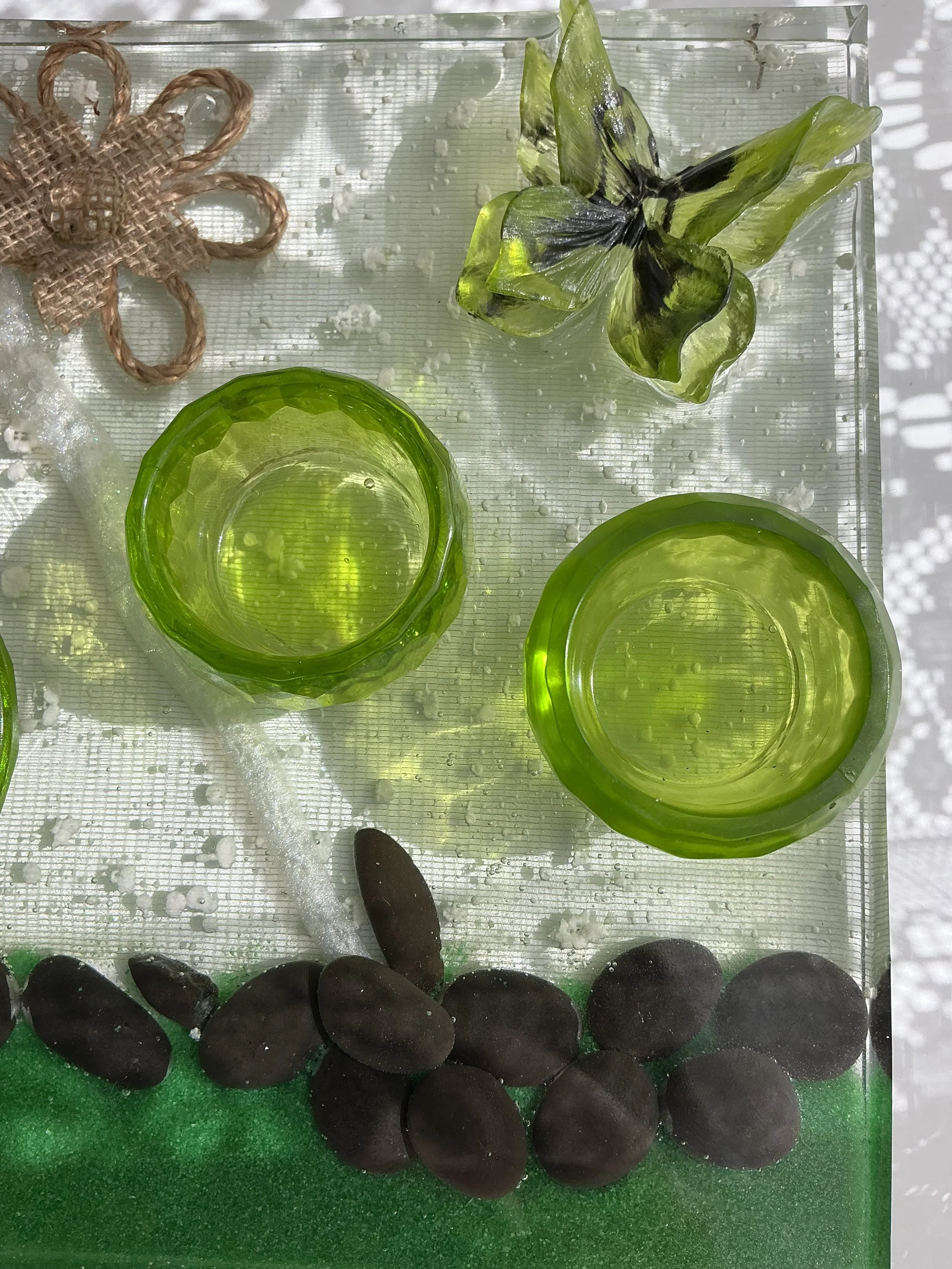 Close-up of the translucent green butterfly and two bright green plant holders. Sunlight reveals delicate black butterfly markings, faceted vessel edges, tiny resin bubbles and shimmering details.