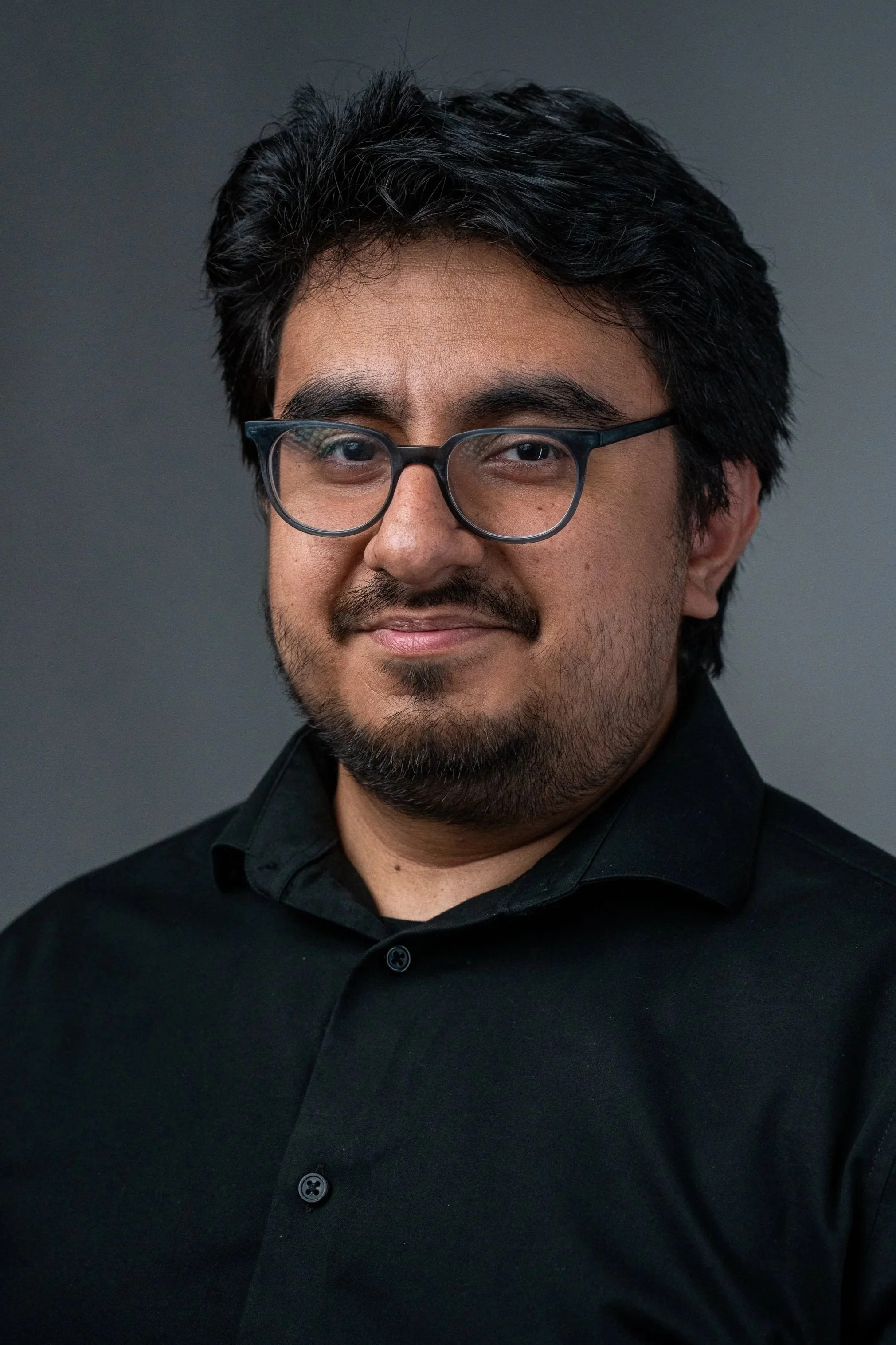 Headshot of a man with glasses and dark hair, wearing a black shirt, smiling slightly, against a gray background.