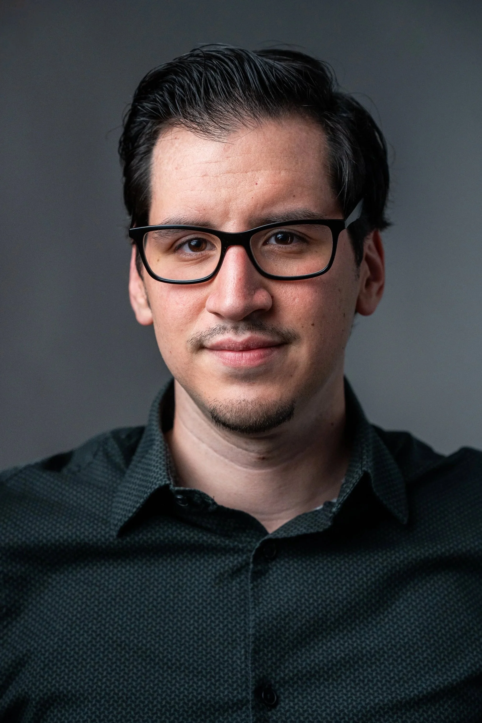 A young man with dark hair, glasses, and a light beard is smiling slightly, wearing a dark button-up shirt, against a dark gray background.