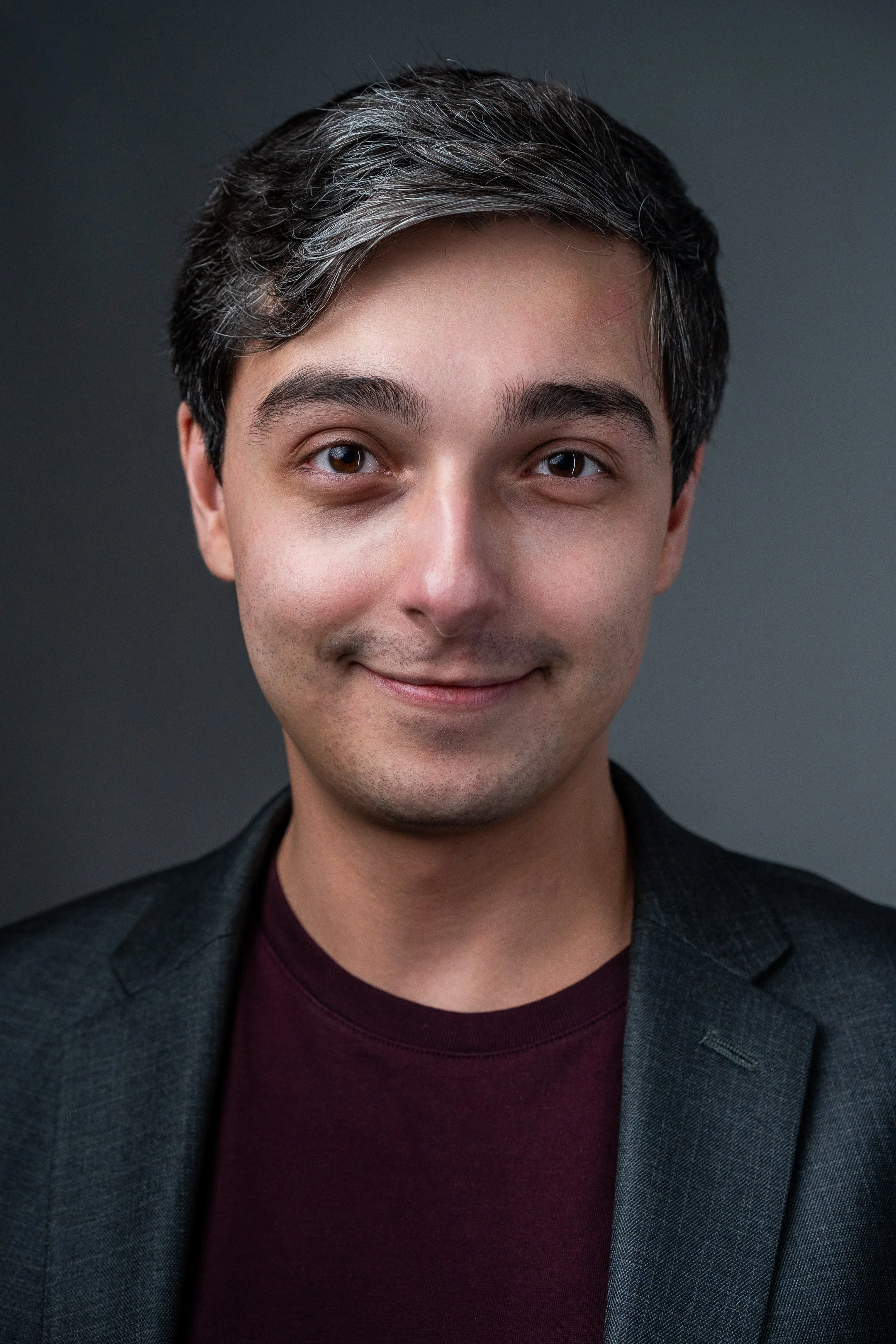 A smiling man wearing glasses, a dark blazer, and a maroon shirt, with a neutral background.