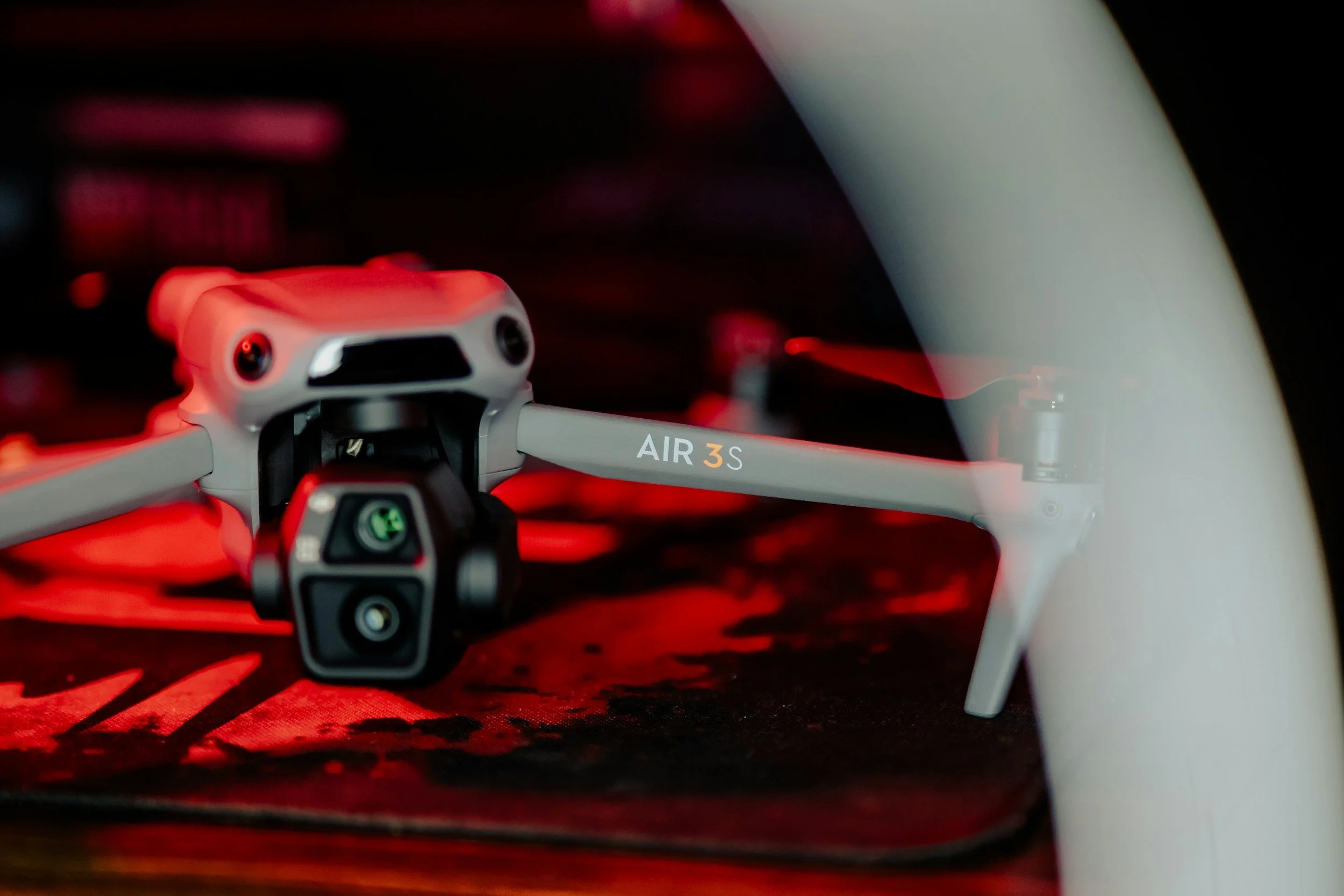 Close-up of a DJI Air 3S drone on a red and black surface, with a white tripod or stand in the background.