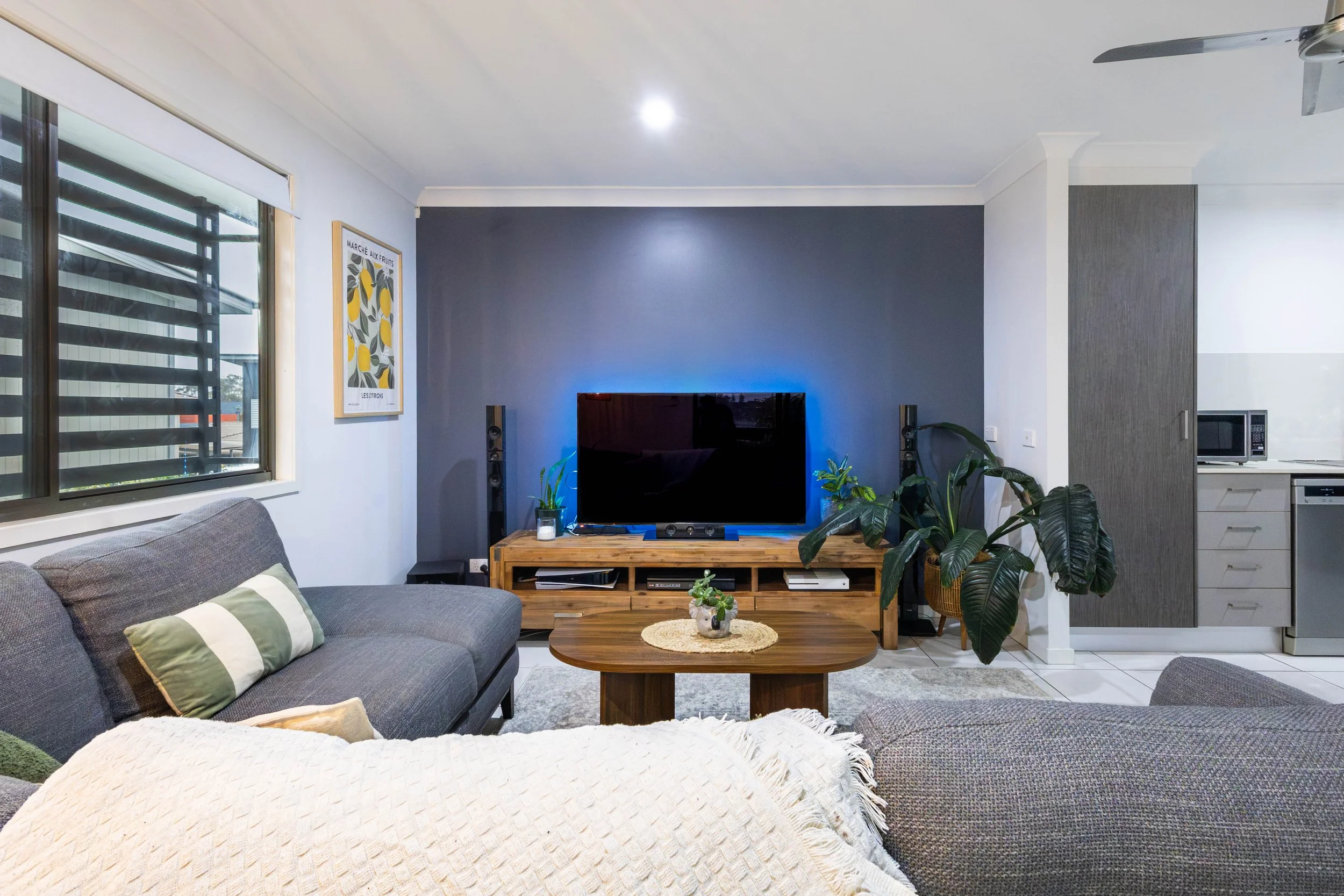 Living room with gray sofa, striped pillows, wooden coffee table with small plant, TV on a wooden stand, blue accent wall, plants, window with blinds, framed artwork, and kitchen area to the right.