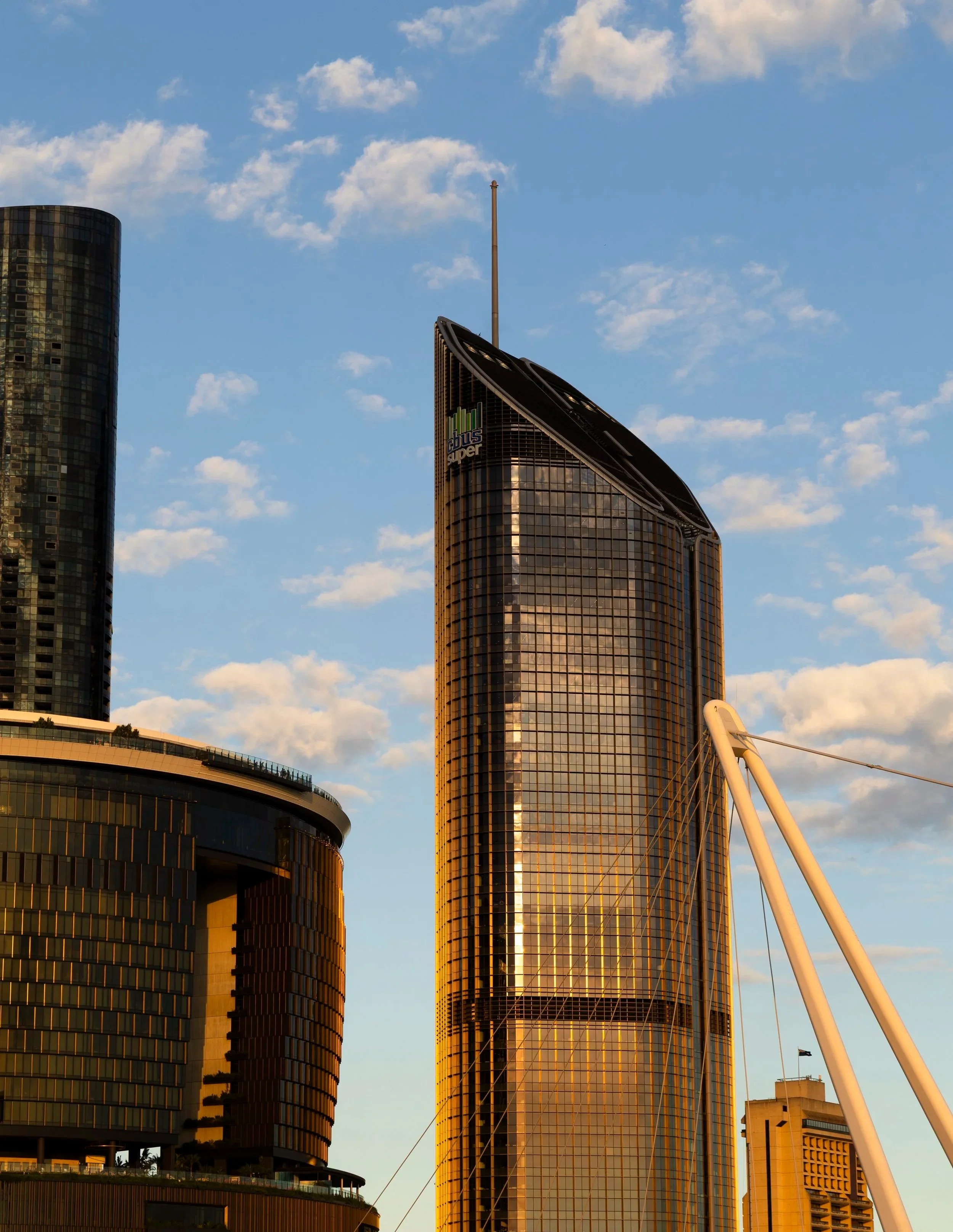 Tall modern skyscraper with glass exterior reflecting the sunset and a blue sky with scattered clouds. Nearby building with curved design and dark windows. Structure with a white arch in the foreground.
