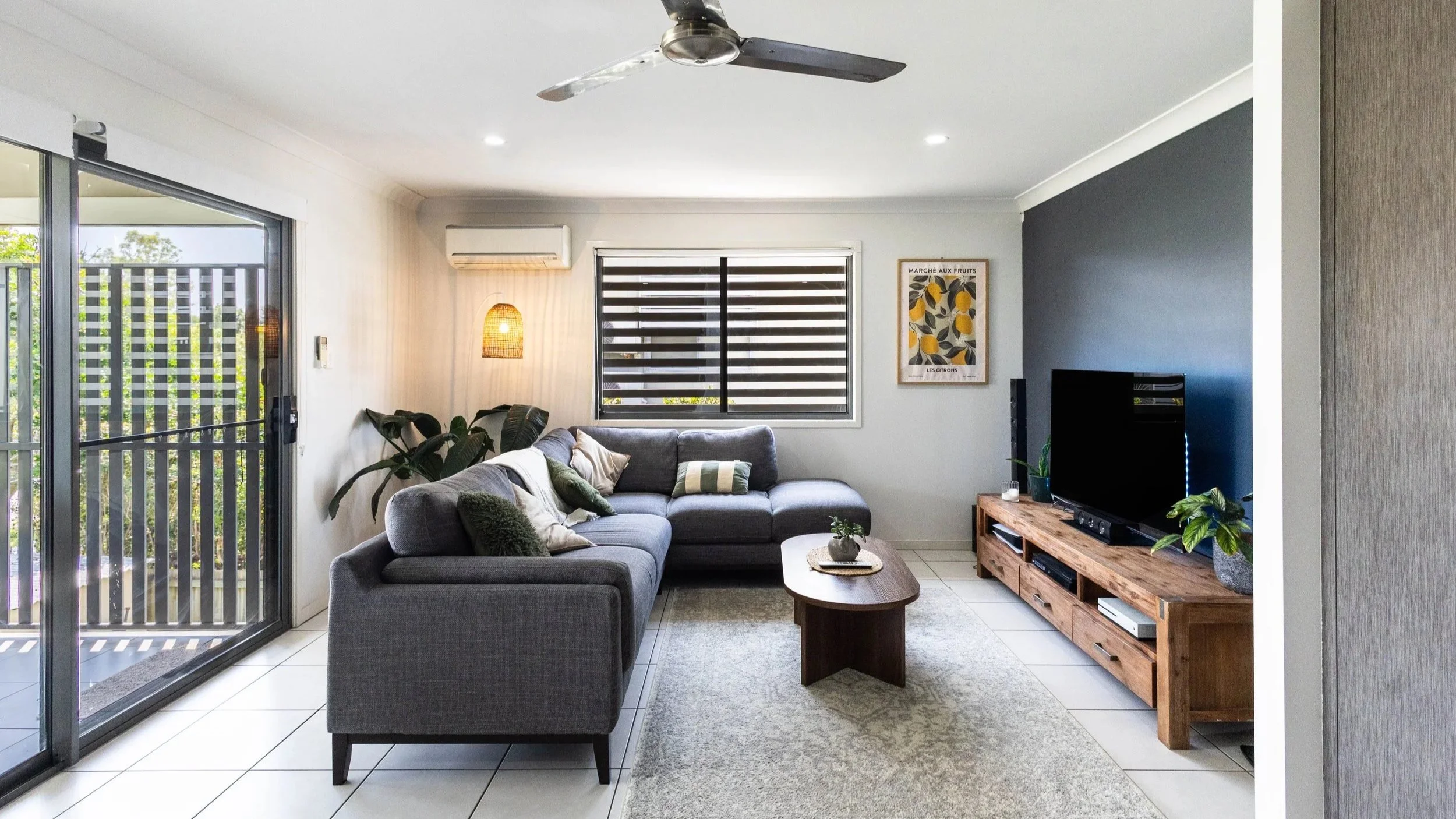 Living room with gray sectional sofa, wooden coffee table, TV on wooden stand, sliding glass door leading outside, window with blinds, indoor plants, framed artwork, ceiling fan, air conditioning unit, and a rug.