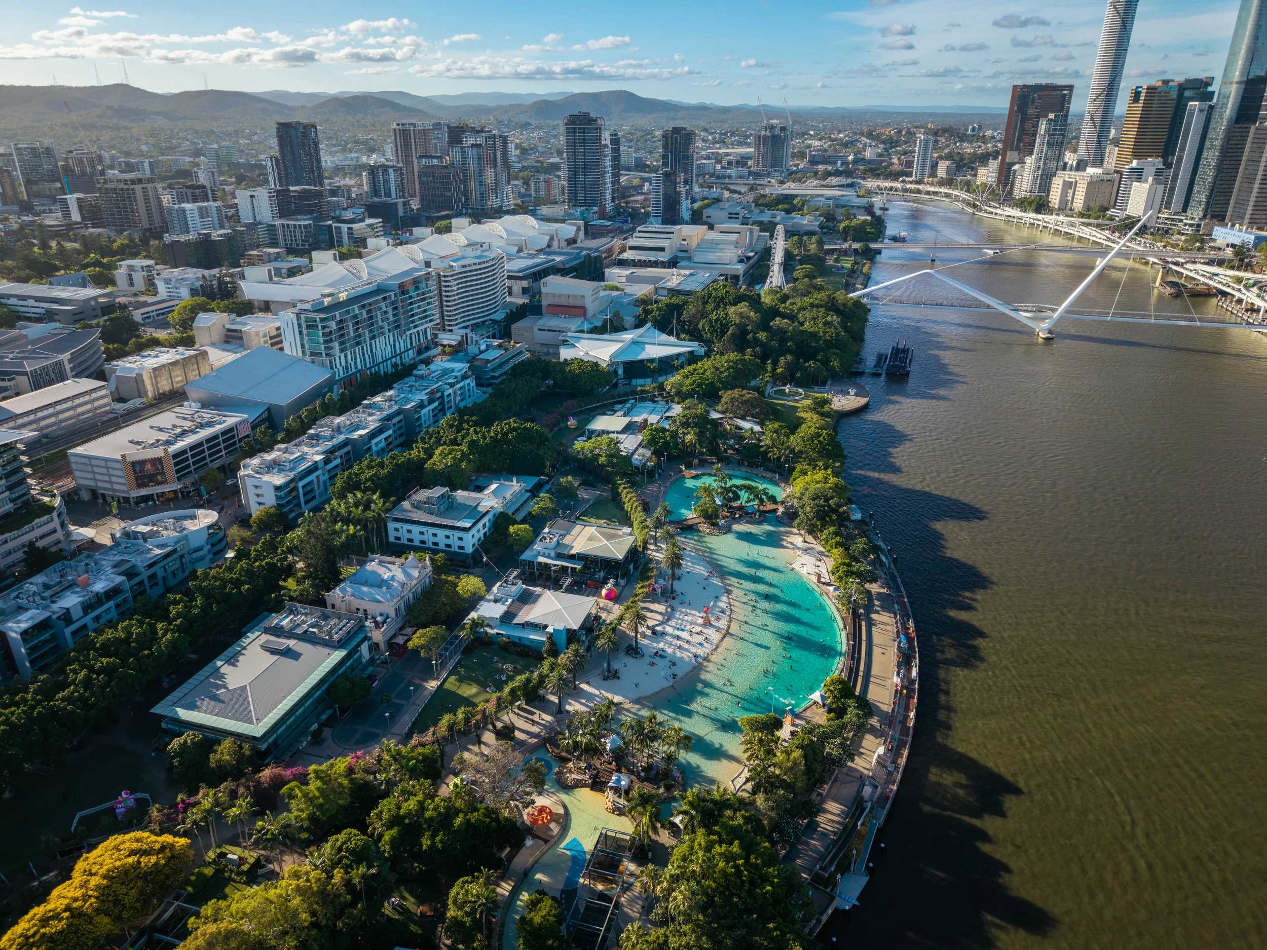 Aerial view of Brisbane city with skyscrapers, river, green parks, and a large outdoor pool area.