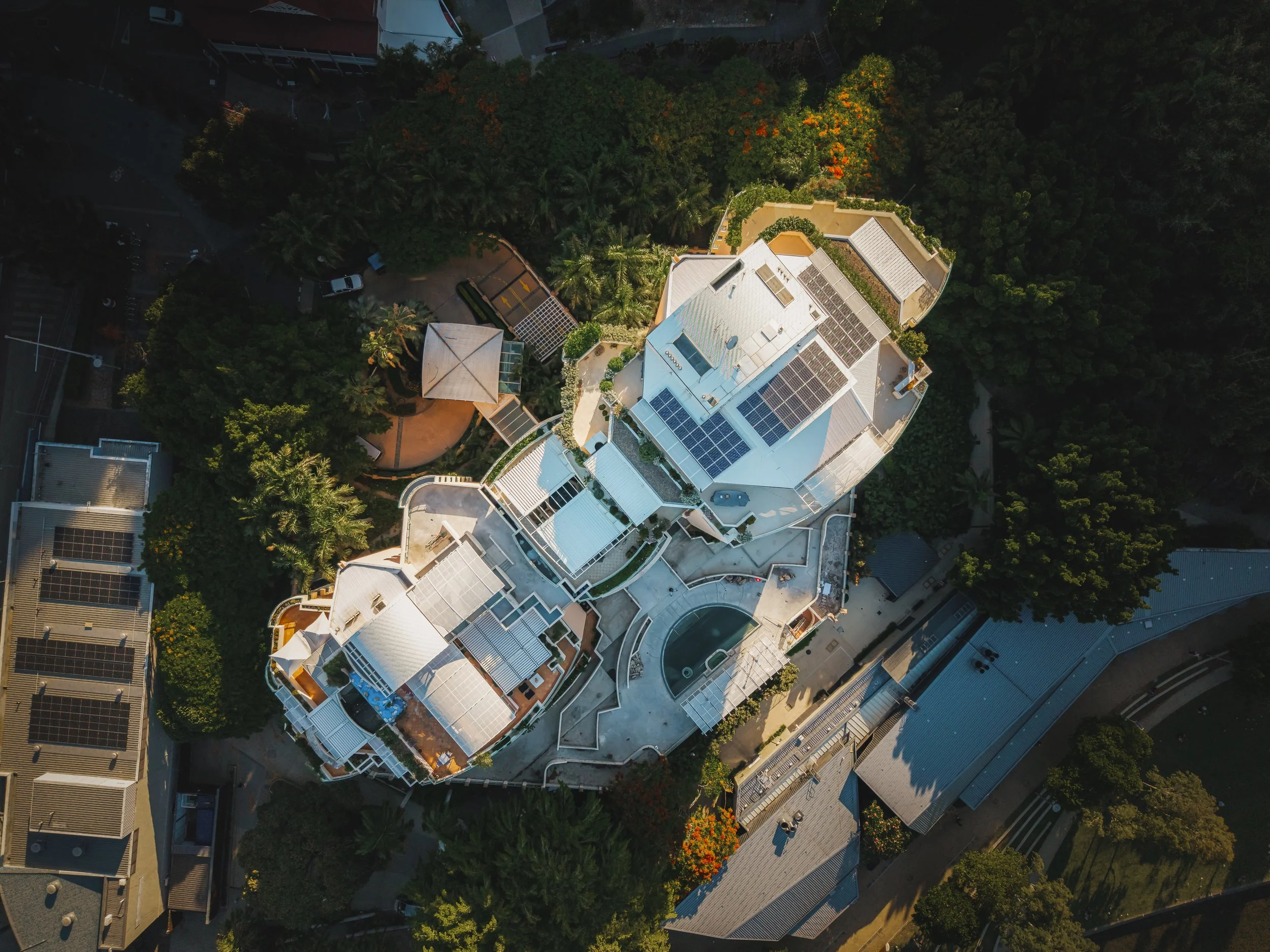 An aerial view of a large, white multi-story house with solar panels on the roof, surrounded by greenery, trees, and other buildings in a neighborhood.
