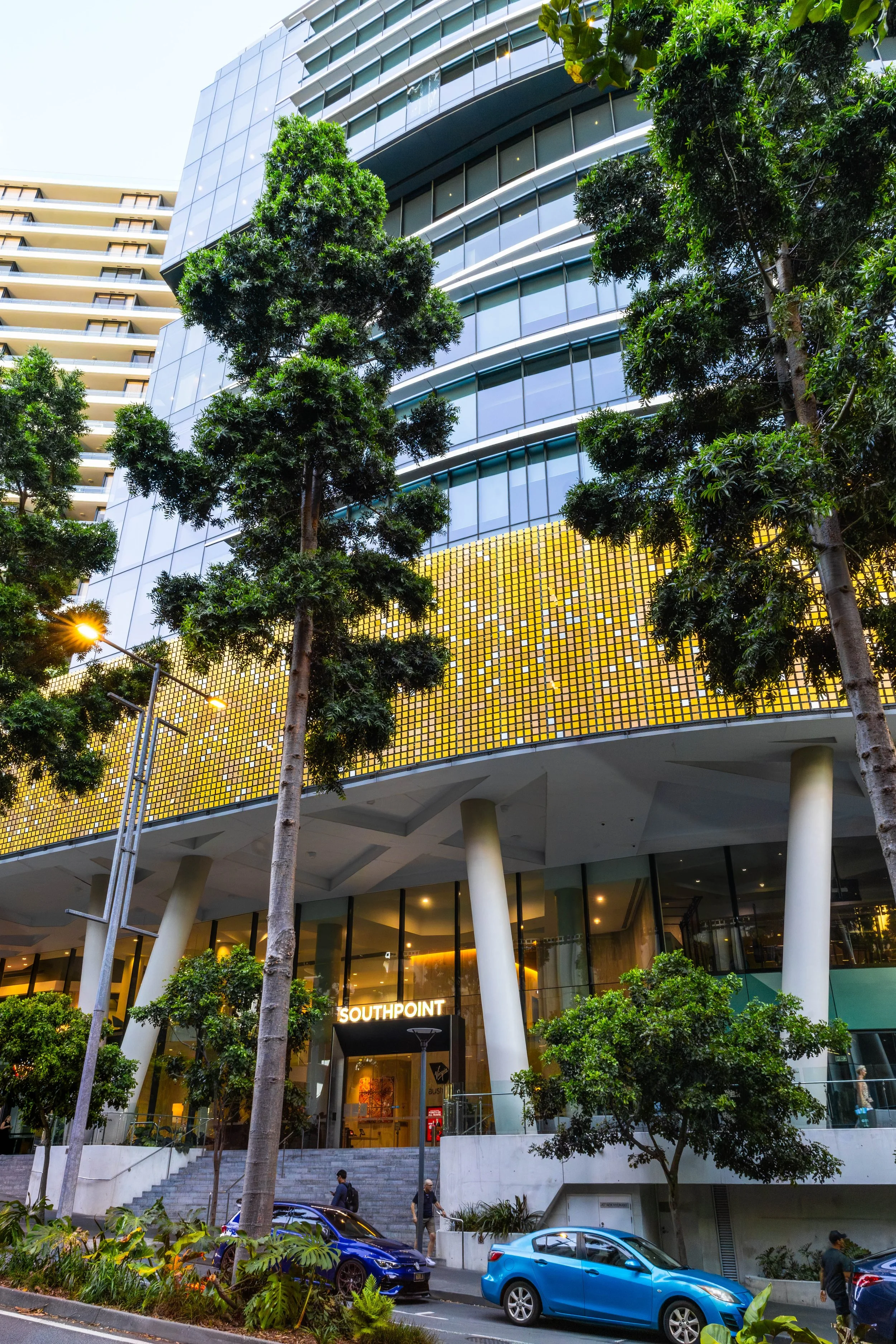 Modern multi-story building with glass and gold facade, surrounded by green trees and parked cars, with pedestrians in front of the entrance labeled "Southpoint".