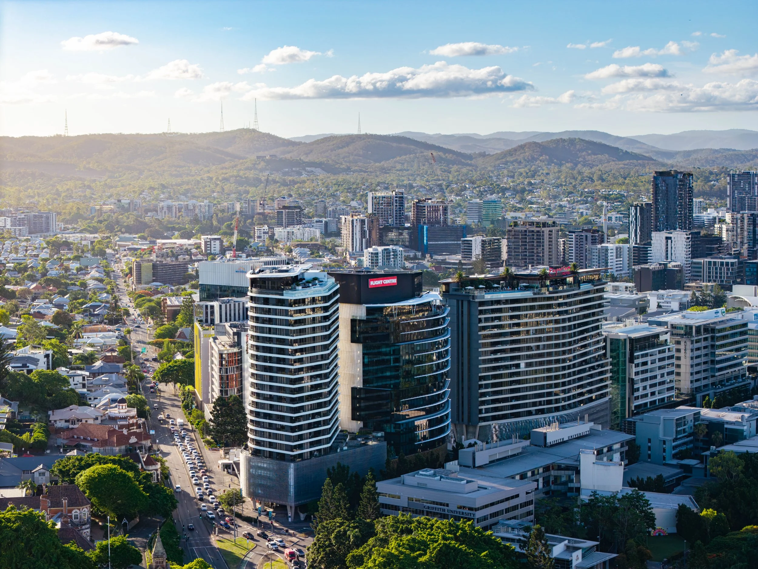 A cityscape with modern high-rise buildings in the foreground and rolling hills with communication towers in the background under a partly cloudy sky.