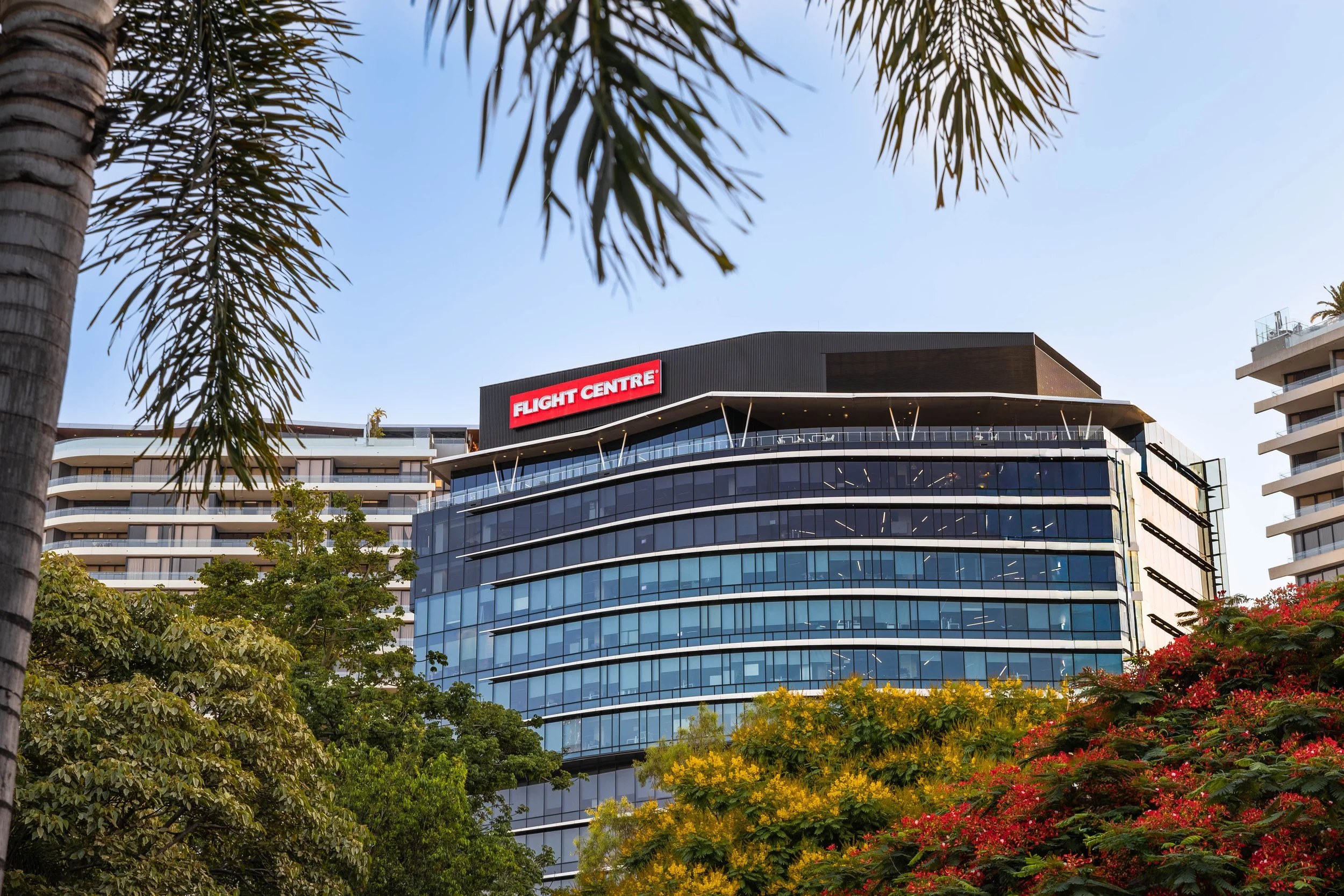 Modern multi-story glass building with a sign that reads 'Flight Centre' at the top, surrounded by trees and foliage, with a clear blue sky overhead.