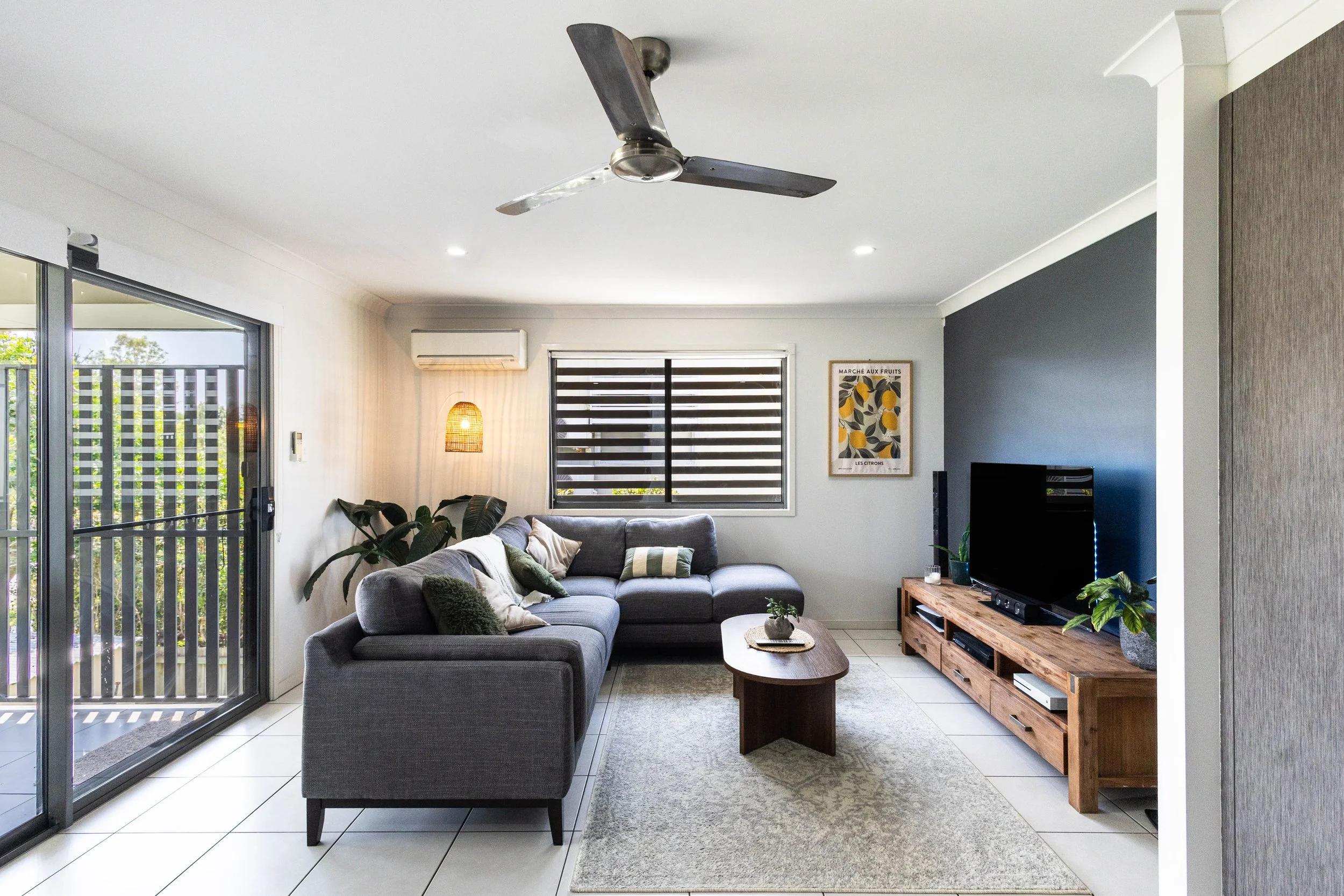 Living room with gray sectional sofa, wooden coffee table, modern tv stand with TV, potted plants, window with blinds, ceiling fan, air conditioning, framed art, sliding glass door leading outside, and a rug on tiled floor.