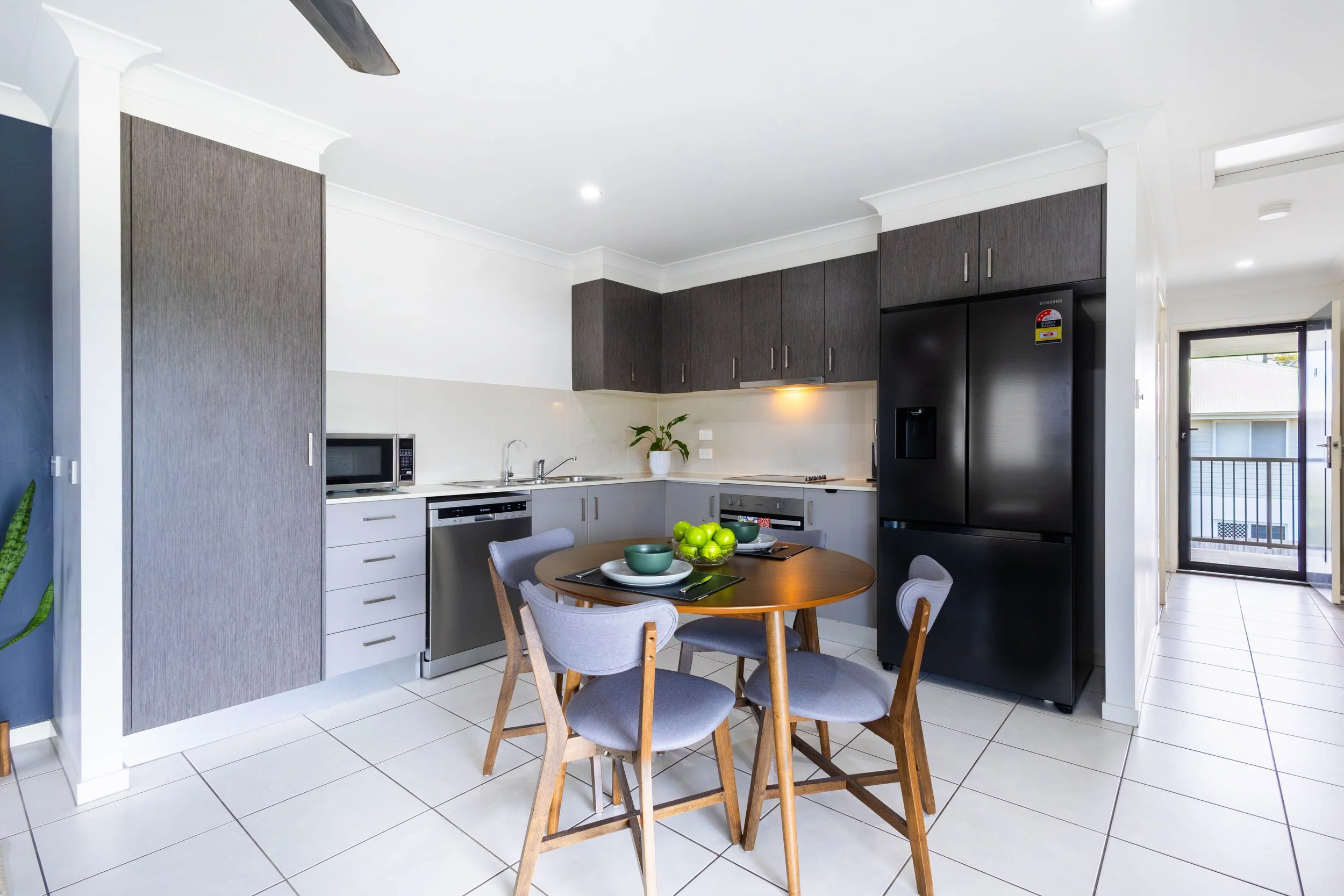 Modern kitchen with gray and white cabinetry, round wooden dining table with chairs, black refrigerator, small window, and a potted plant.