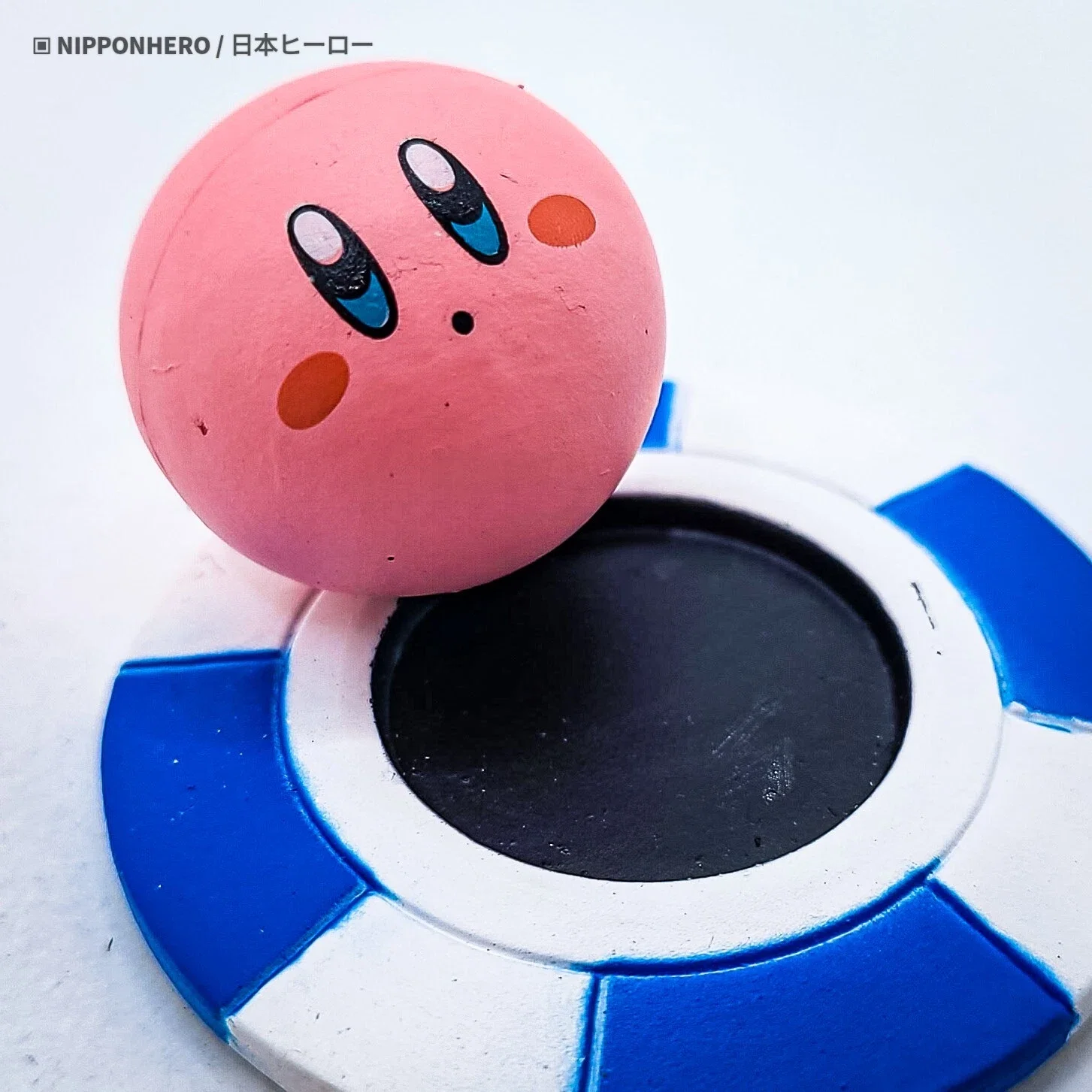 A pink spherical object with a face drawing resembling Kirby from video games, positioned on a circular black and white base with blue segments.