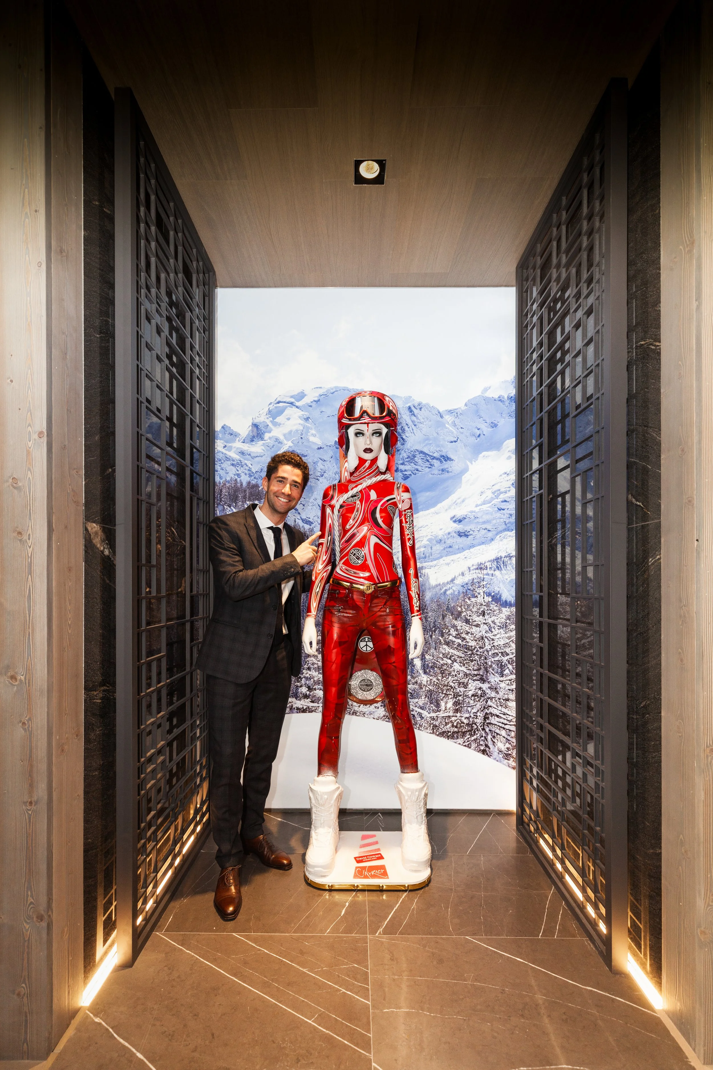 Un homme en costume noir souriant, pointant vers une mannequin toute en rouge avec un casque et des bottes blanches, dans une salle avec un décor de montagnes enneigées en arrière-plan.