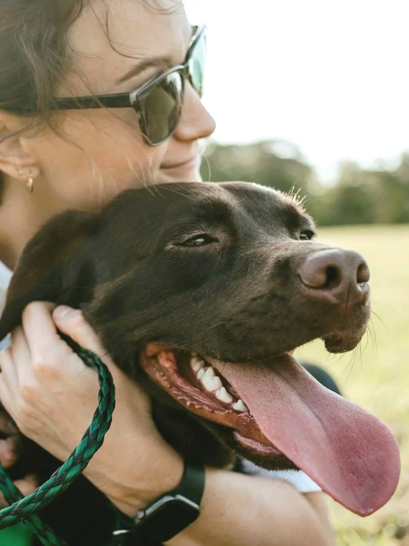 A woman wearing sunglasses and earrings gently holding a happy black dog with a wide open mouth and tongue hanging out outdoors.
