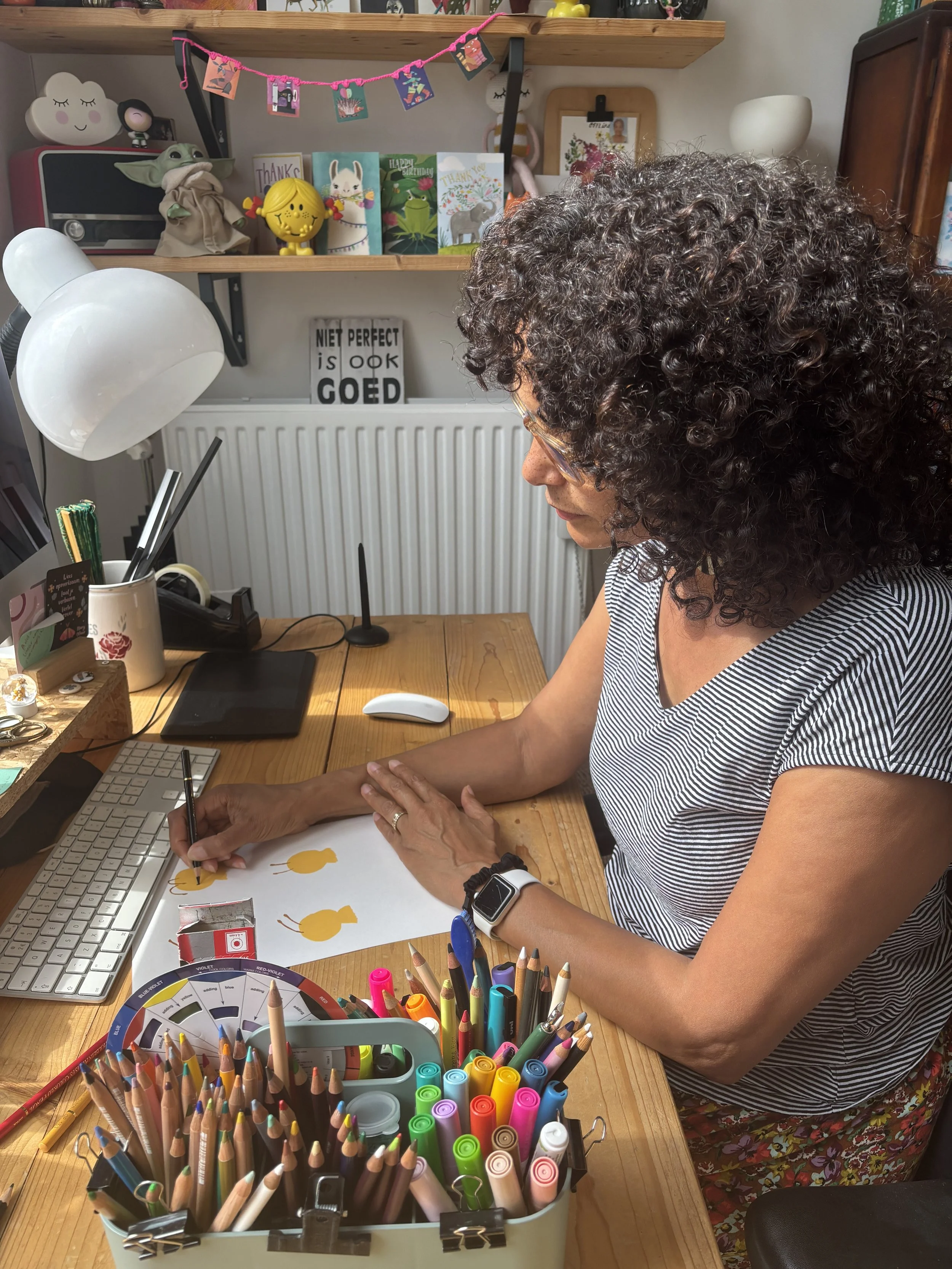 Maruga is sitting at a wooden desk, drawing shapes on a white sheet of paper. The desk has a keyboard, a computer mouse, and various colored pens and markers. Behind her, there are shelves with greeting cards and decorative items