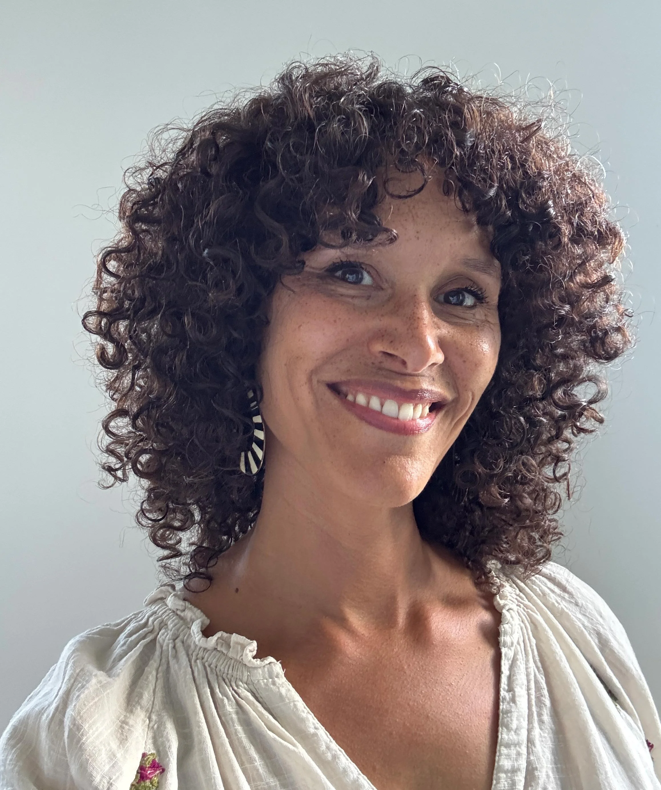 A woman with curly brown hair smiling at the camera, wearing a white embroidered blouse, against a light gray background.