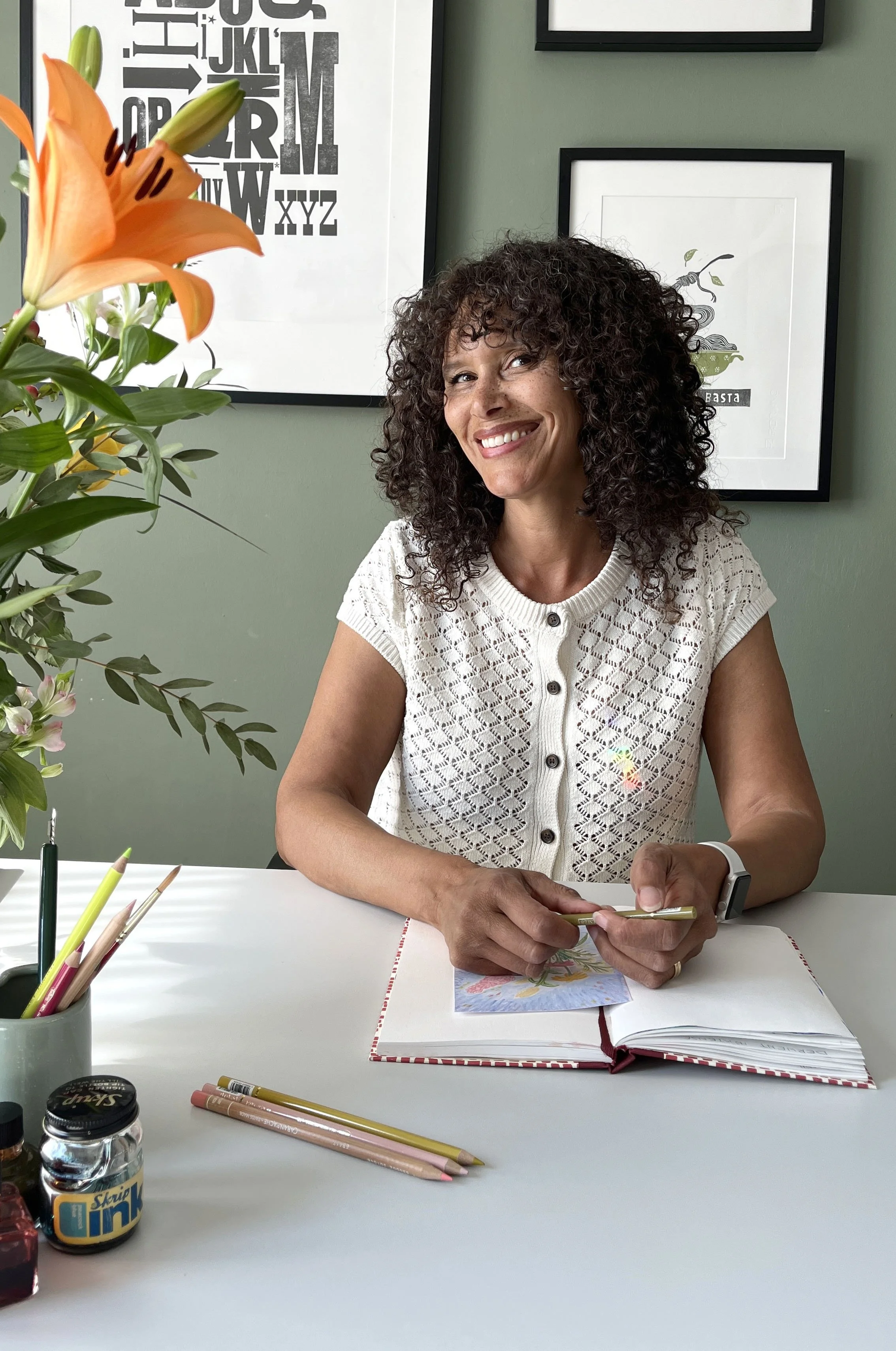 Maruga Koops, smiling and sitting at a white table, holding a gold pen. The table has art supplies, including colored pencils, a sketchbook, and a jar of ink.