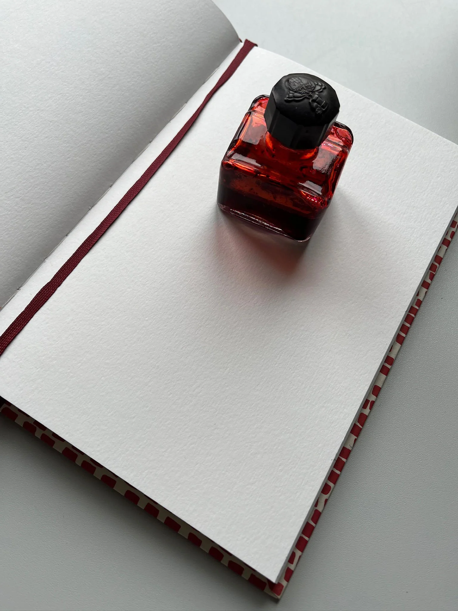 A red ink bottle with a black screw cap placed on a white textured surface, oriented diagonally from the bottom left to the top right, with a red cloth-bound notebook underneath.