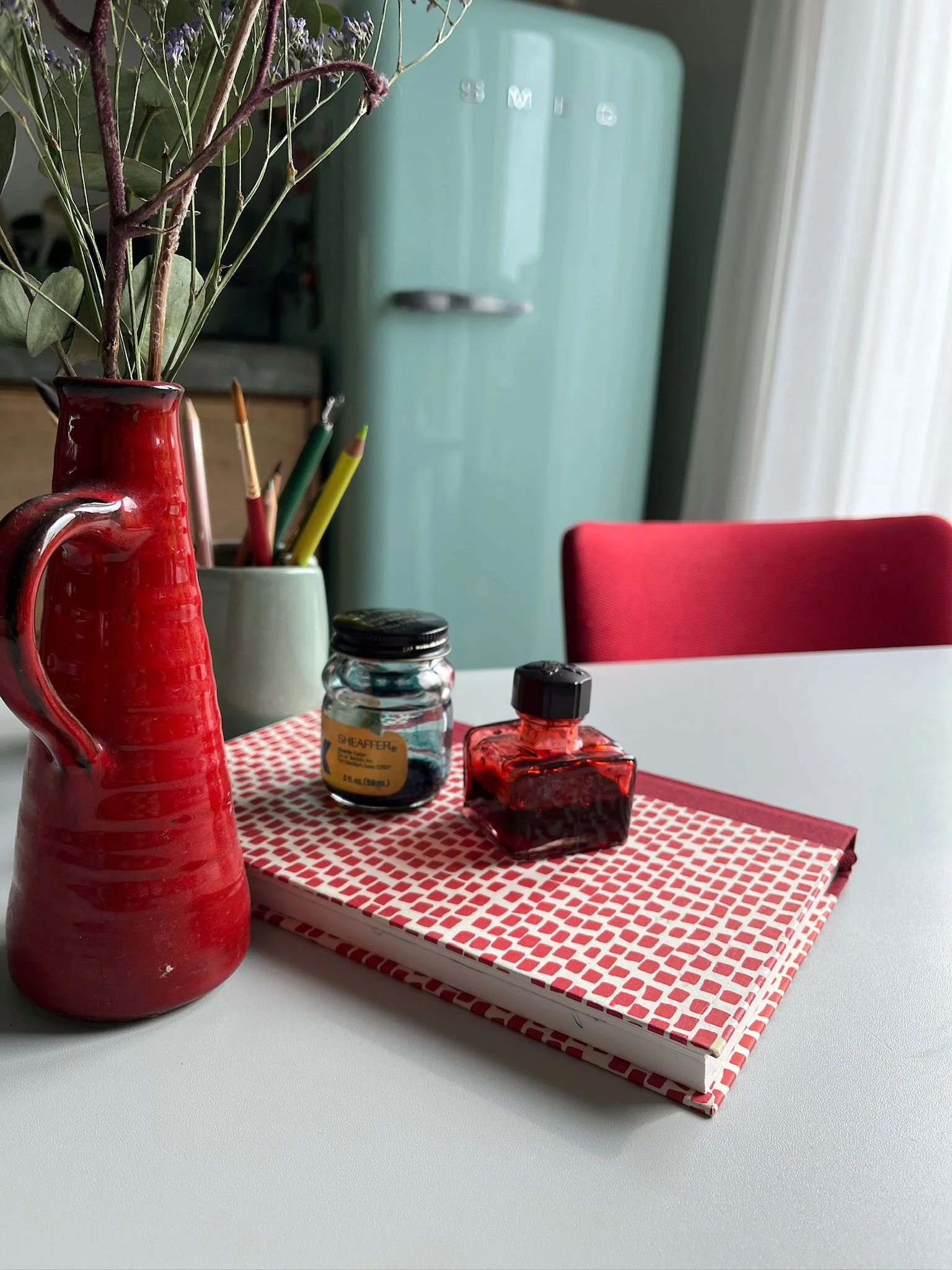 Decorative red pitcher holding dried purple flowers and green leaves, two ink bottles, and a set of colorful pens on a red and white checkered napkin on a white table.
