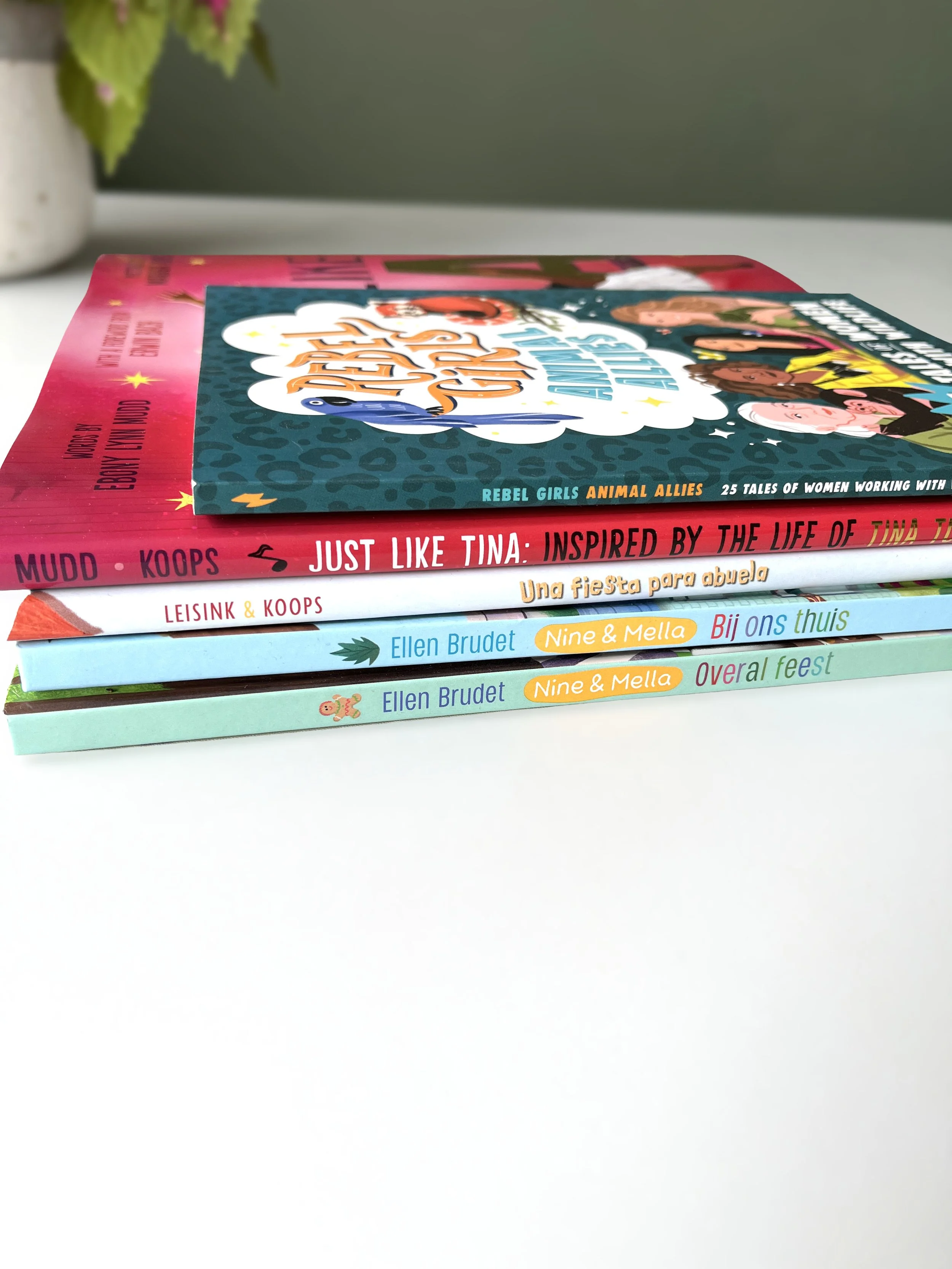 A stack of several colorful children's books with titles such as 'Just Like Tina' on top, placed on a white surface near a potted plant.