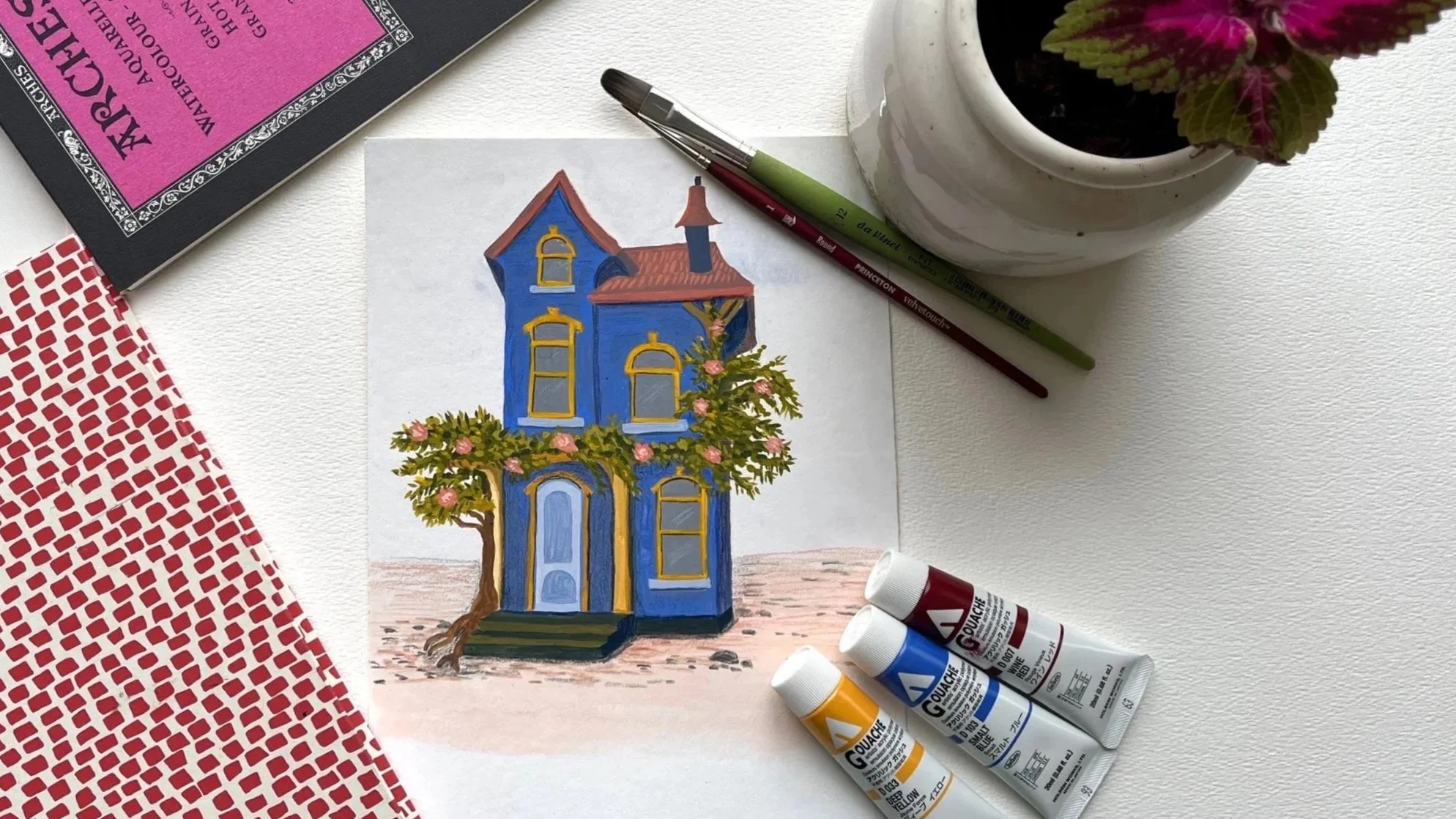 A colorful illustration of a blue house with yellow window frames and a red roof is on a white sheet of paper, surrounded by paints, brushes, a potted plant, and a book on a white surface.