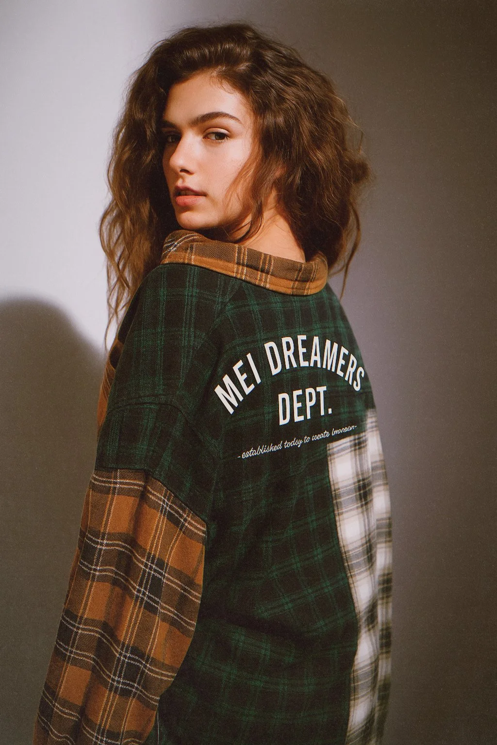 Young woman with wavy brown hair wearing a patchwork plaid shirt with text on the back that reads 'MEI DREAMERS DEPT.'
