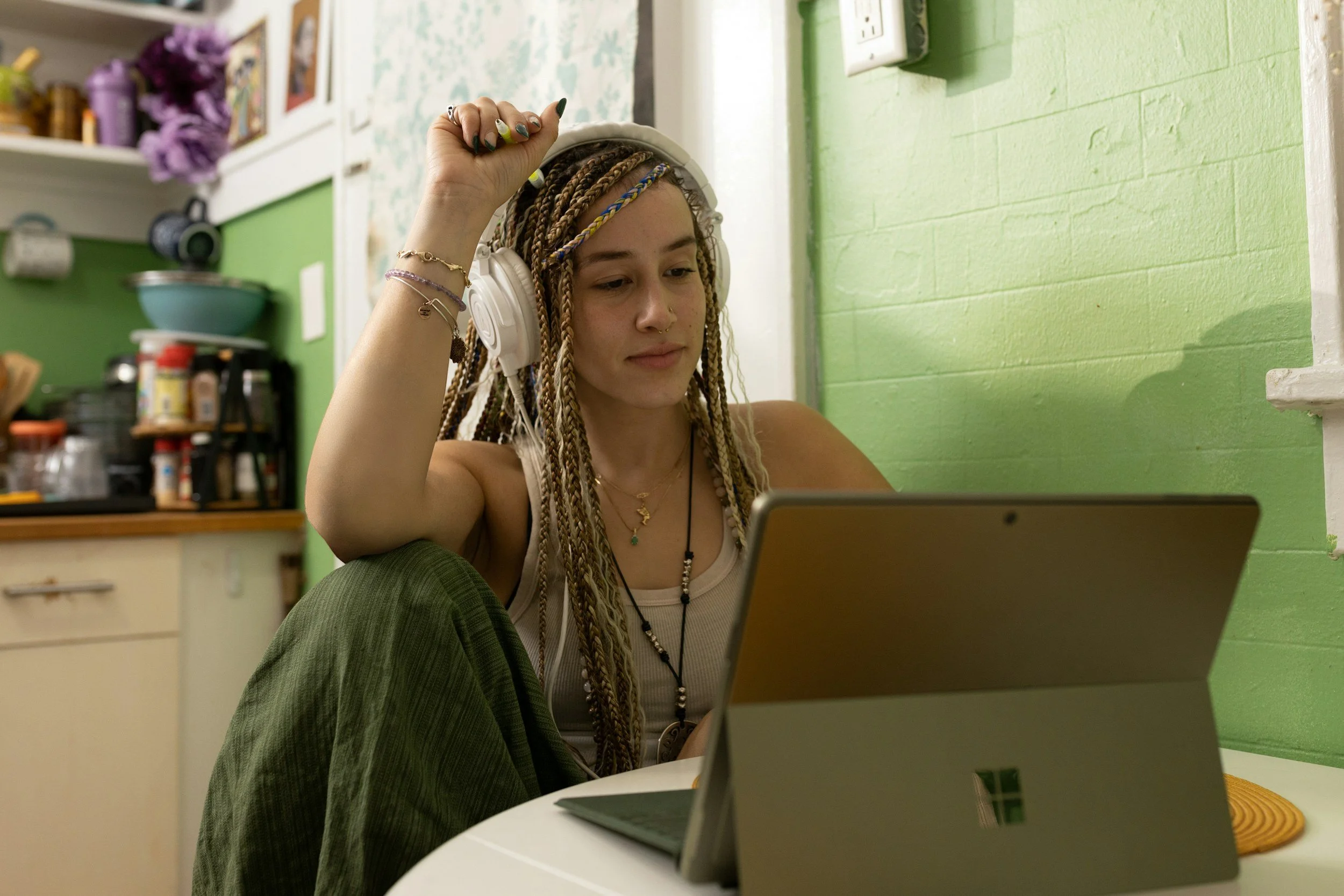 A young woman with long braided hair, wearing headphones and a tank top, is sitting at a white table using a Microsoft Surface tablet. She appears relaxed, with a green wall behind her and a kitchen in the background.