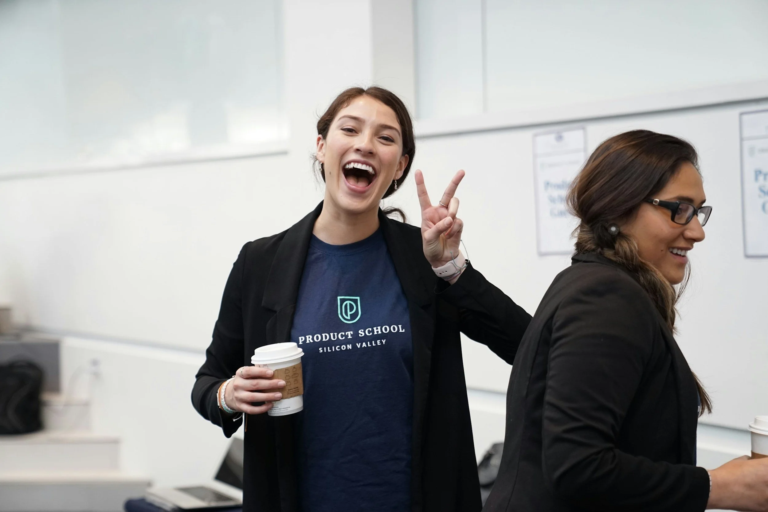 Two women smiling and holding coffee cups, standing in a classroom or training room.
