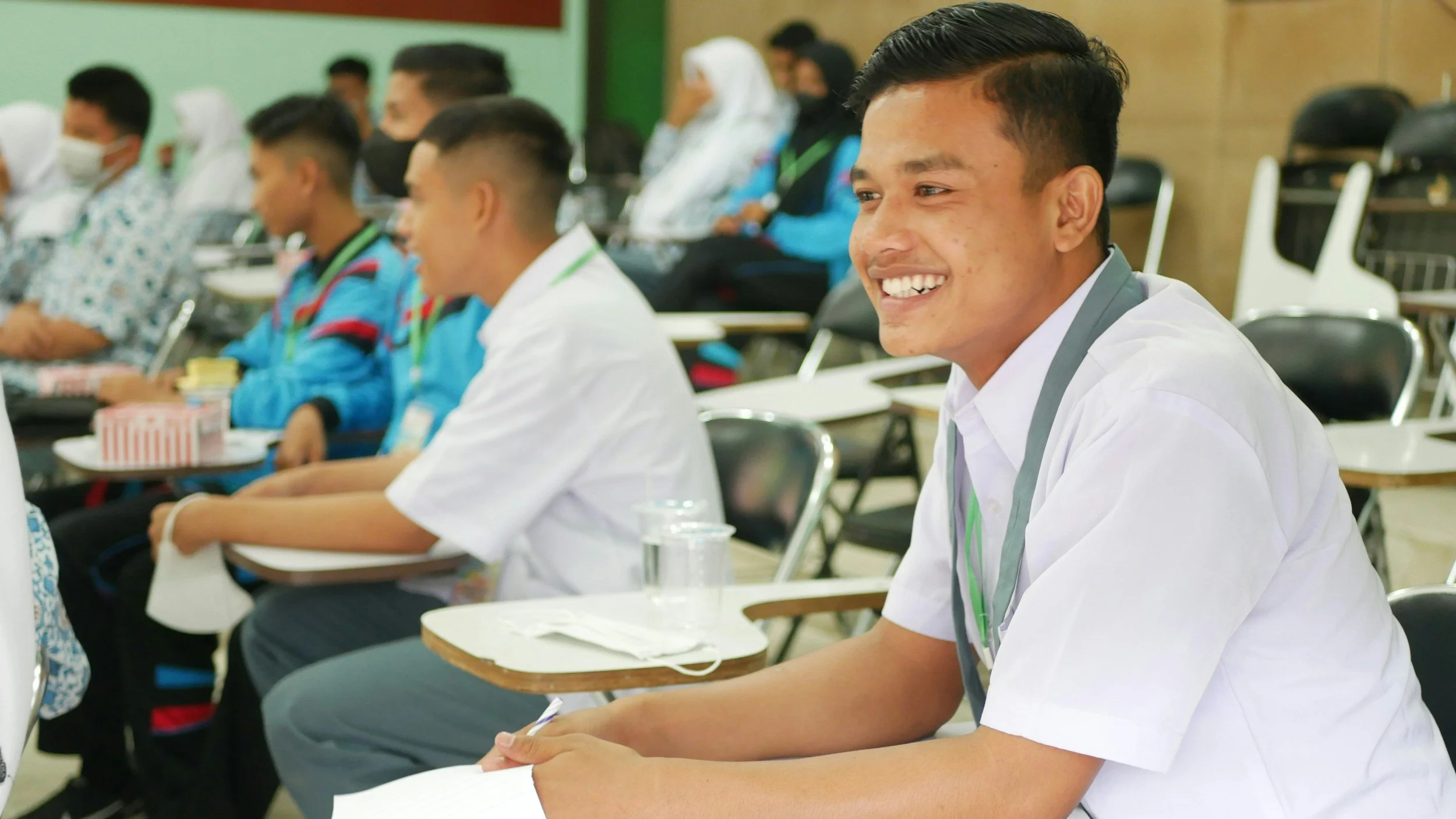Smiling student in white shirt attending a classroom or lecture hall with other students, some wearing face masks, seated at desks.
