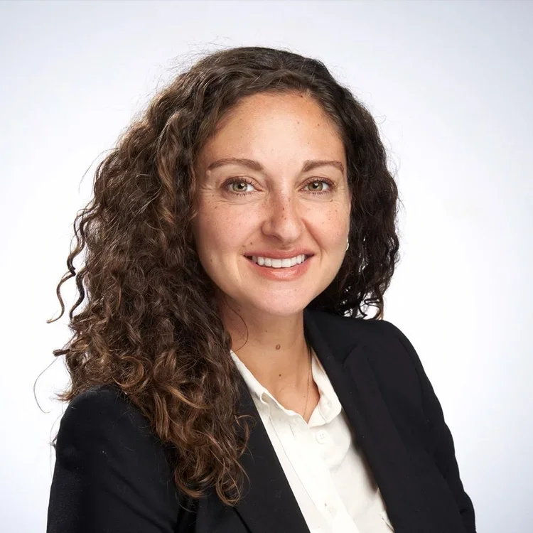 A woman with curly brown hair, smiling, wearing a black blazer and white shirt against a light background.