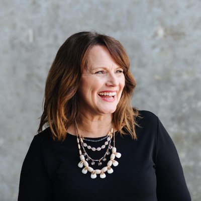 Kim McManus with shoulder-length brown hair, smiling, wearing a black top and layered pearl necklace, against a gray textured background.