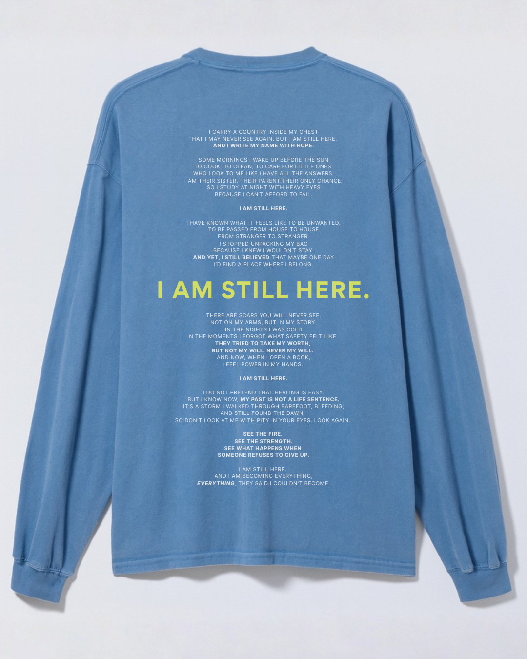 Blue long-sleeve shirt with text printed on the back, featuring a motivational poem about resilience and hope with a large highlighted phrase "I AM STILL HERE" in yellow.