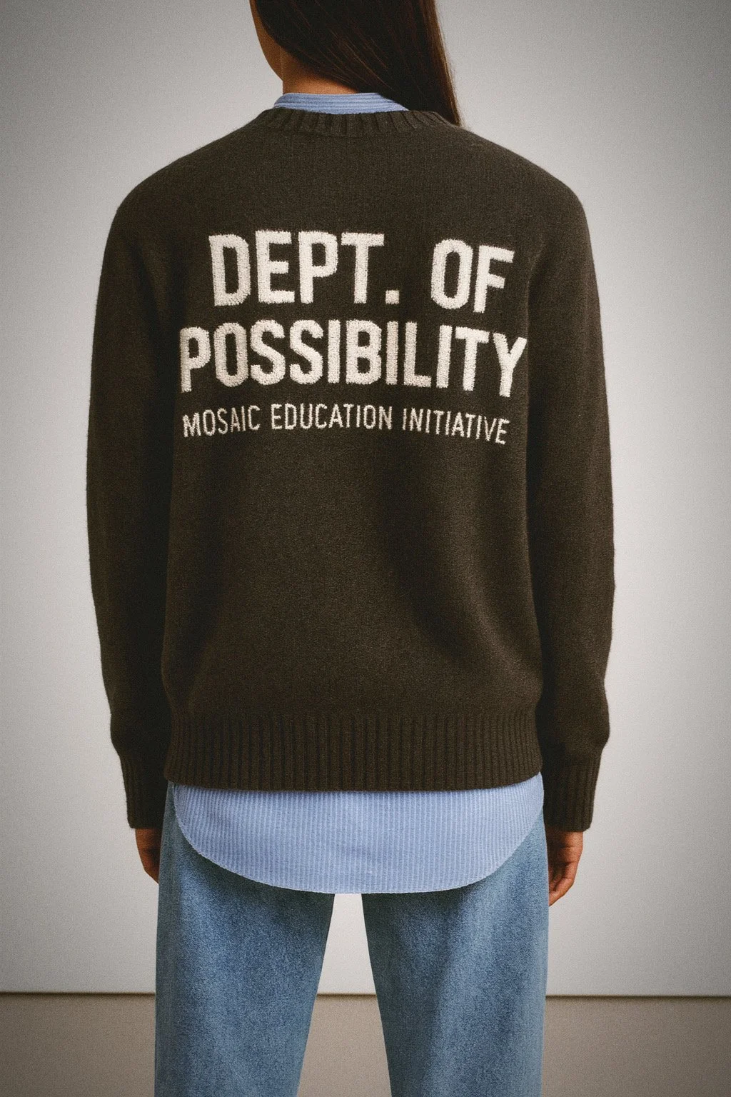 Back view of a person wearing a dark sweater with white text that reads 'DEPT. OF POSSIBILITY' and smaller text 'MOSAIC EDUCATION INITIATIVE', with a blue shirt underneath and light blue jeans.
