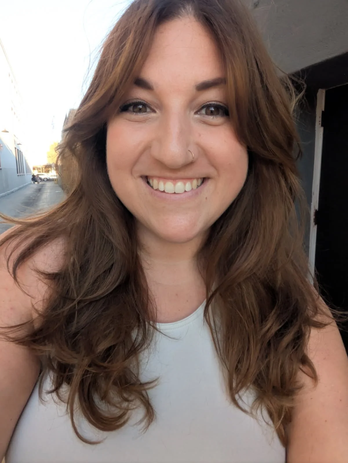 A woman taking a selfie outdoors, smiling with long, curly brown hair, wearing a white top.