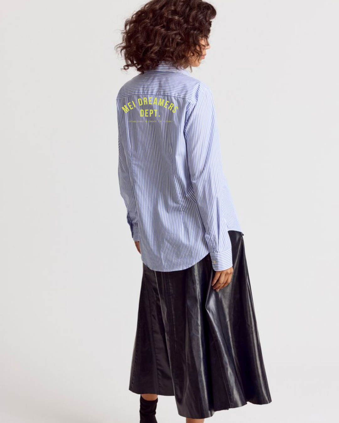 Back view of a woman with curly hair wearing a light blue striped shirt with yellow printed text on the back that says "MEN DREAMERS DEPT." and a long black leather skirt.
