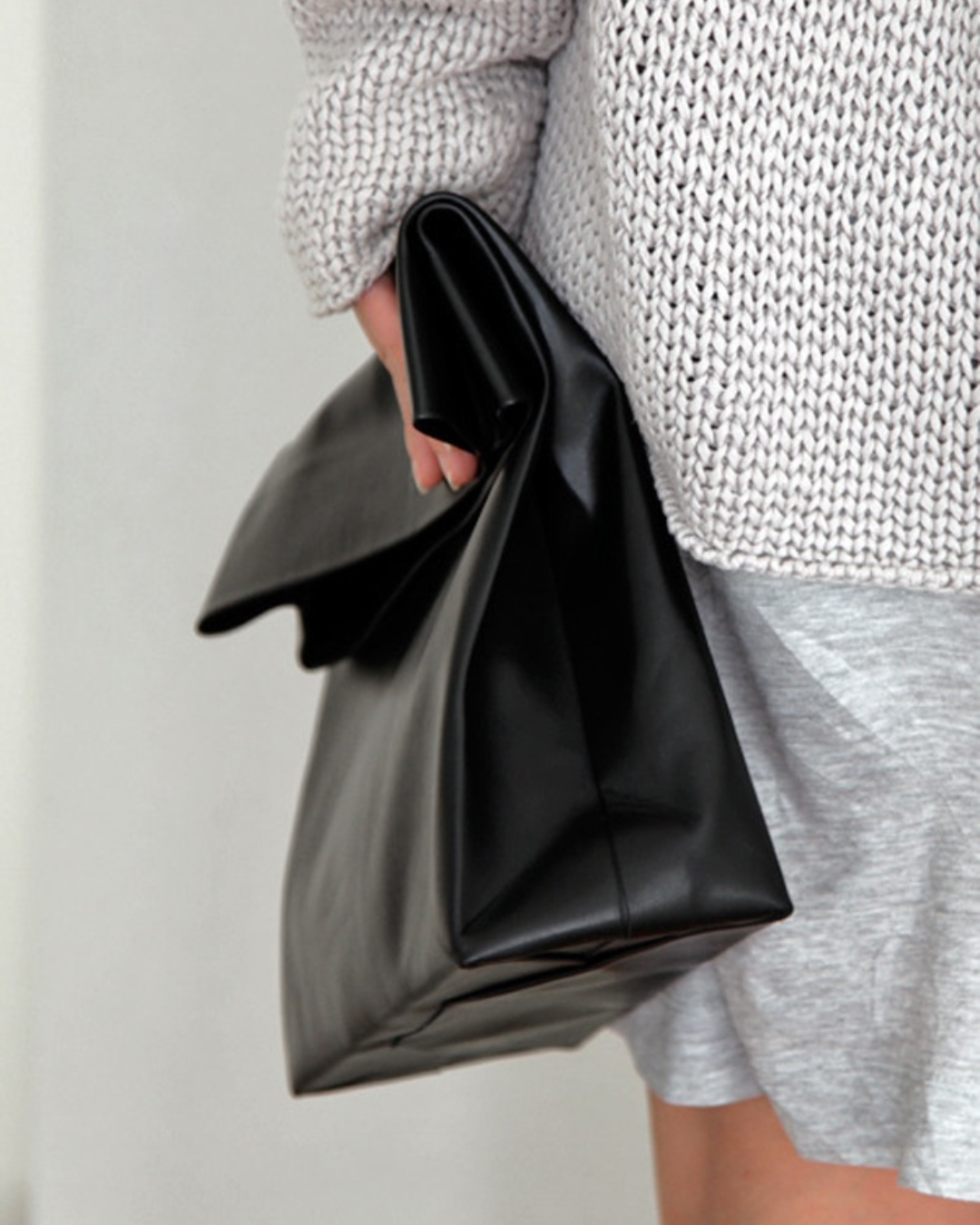 A person wearing a gray knit sweater and gray shorts holding a black patent leather clutch bag.