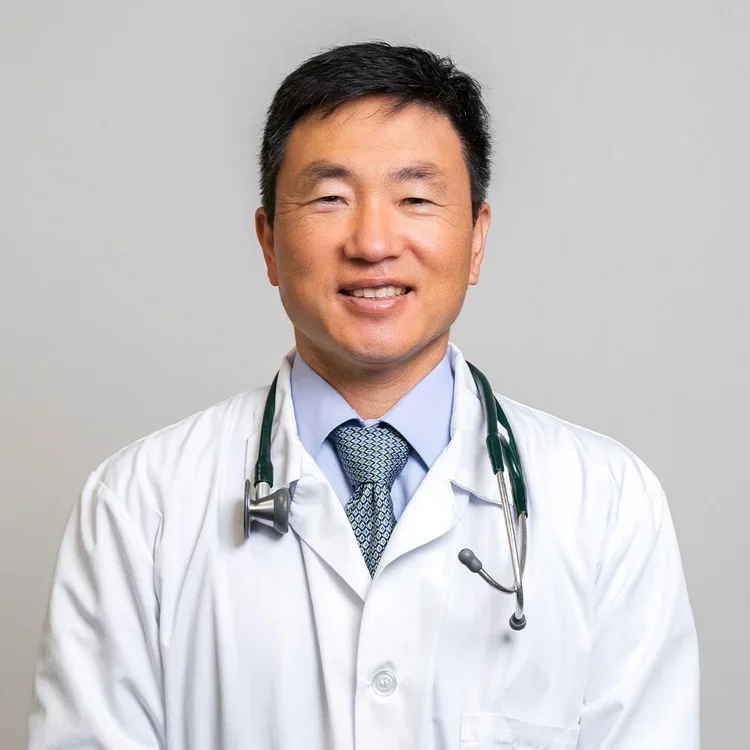 A male doctor wearing a white lab coat, light blue shirt, patterned tie, and a stethoscope around his neck, smiling at the camera against a plain background.