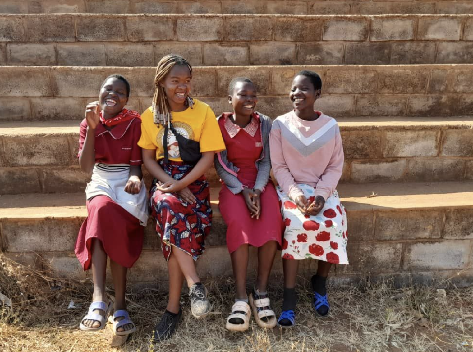 Four girls sitting on stone steps, smiling and laughing, outdoors on a sunny day.