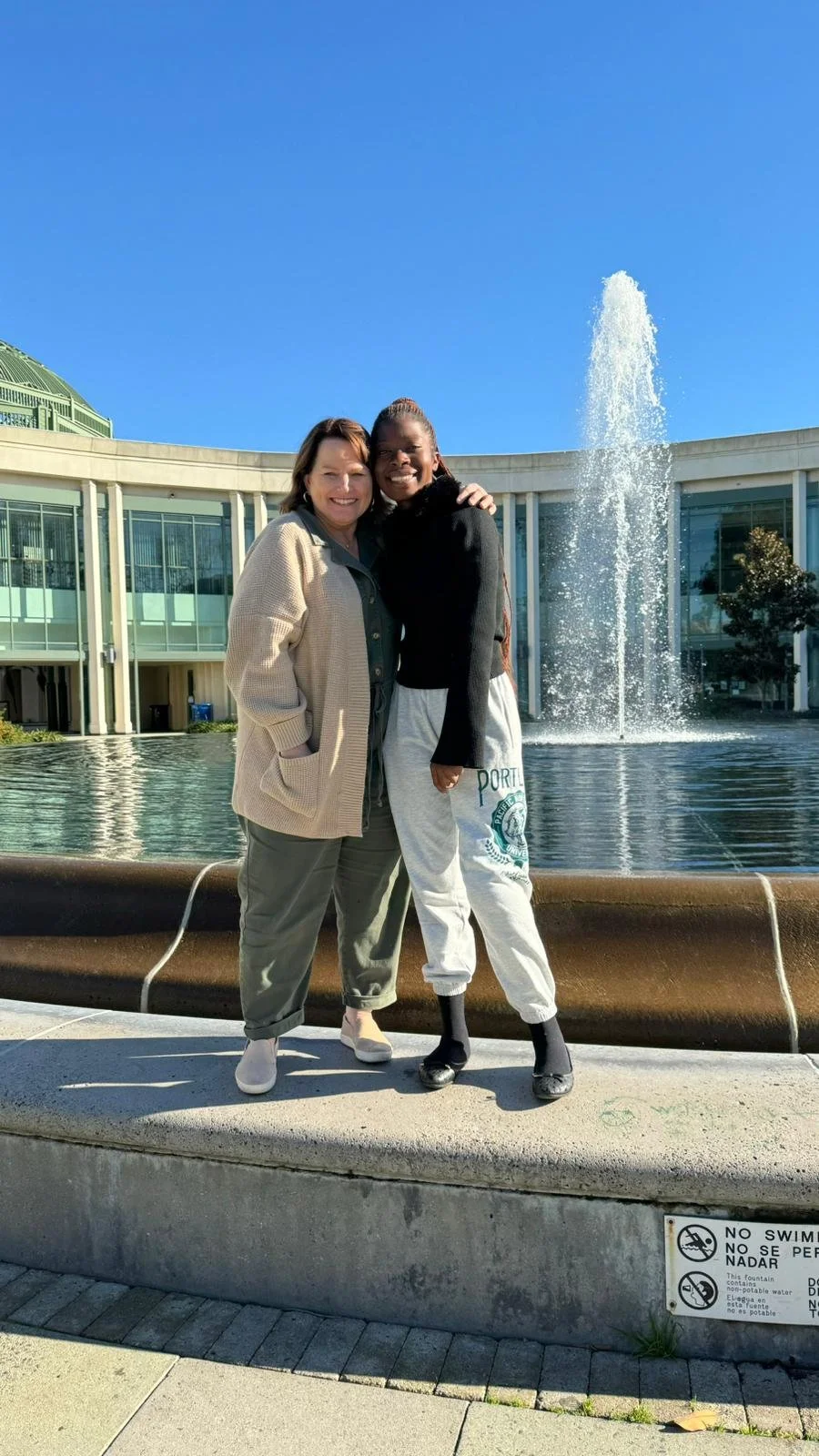 Two women standing together smiling in front of a fountain at the park with a modern building behind them on a sunny day.
