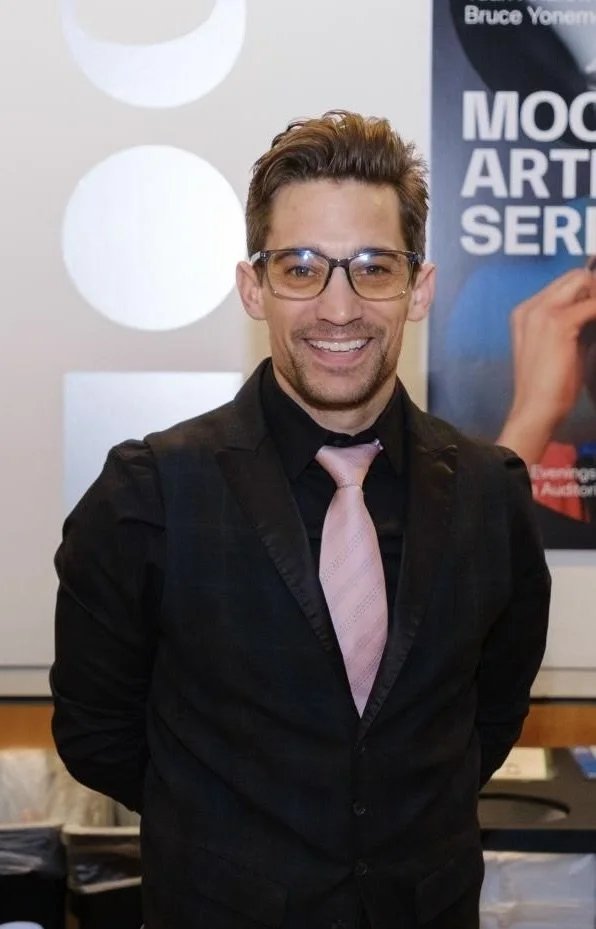 A smiling man with glasses, wearing a black suit with a pink tie, standing indoors in front of a poster and white wall with circular decorations.