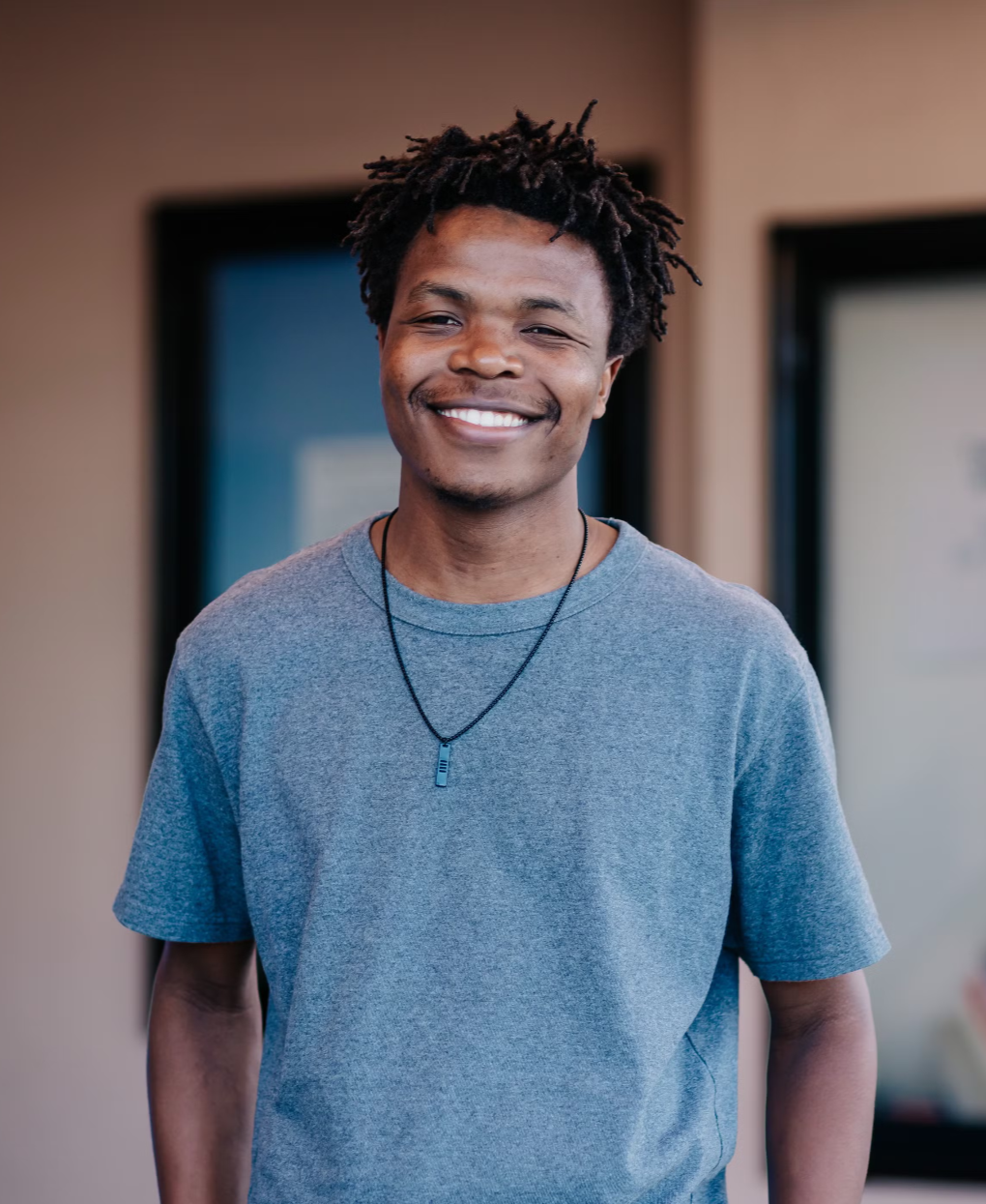 A young man with curly hair smiling, wearing a gray t-shirt and a black necklace, standing indoors.