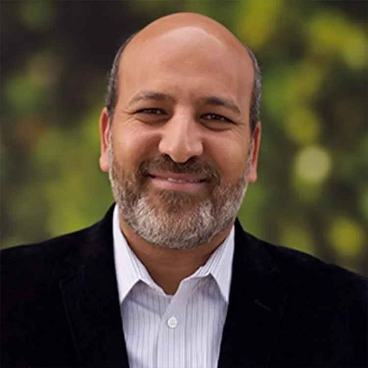 A man with a beard and a friendly smile, wearing a white dress shirt and a black blazer, standing outdoors with a blurred background of greenery.