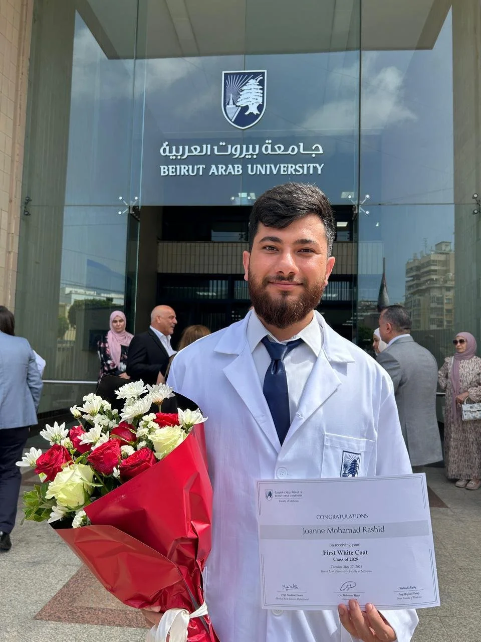 Graduating student in a white coat holding a bouquet of flowers and a certificate.