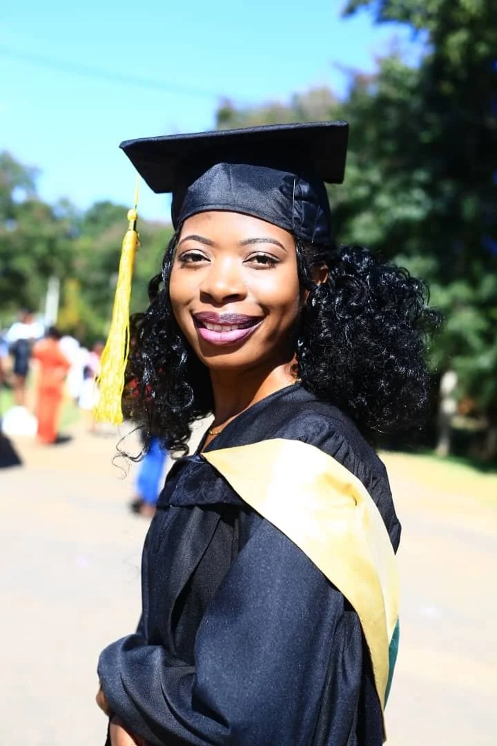 A young woman in a graduation cap and gown outdoors, smiling, with trees and other graduates in the background.