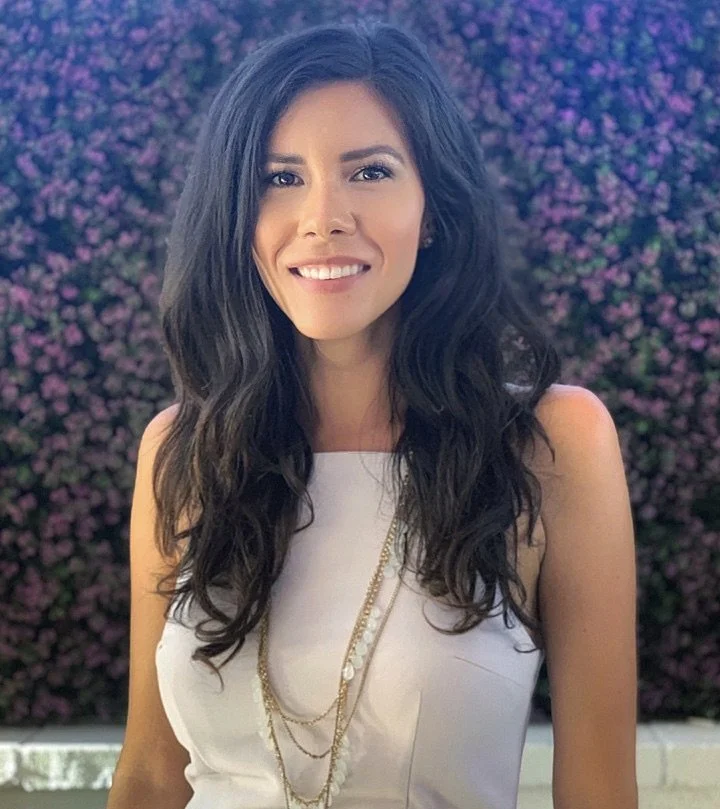 A woman with long wavy dark hair, wearing a white top and layered necklaces, standing outdoors in front of a bush with purple flowers, smiling at the camera.