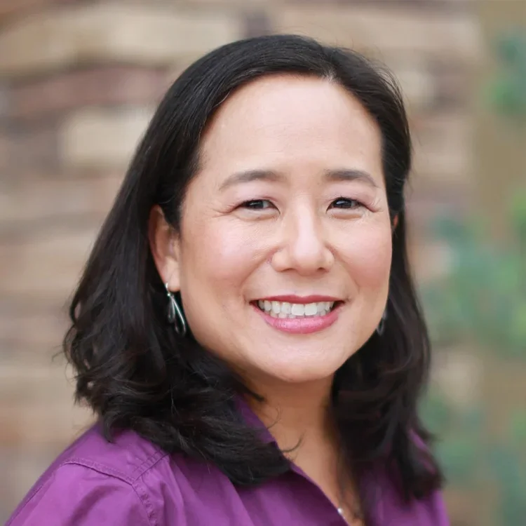 Close-up portrait of a with shoulder-length black hair, wearing a purple shirt and silver earrings, smiling outdoors with a blurred background.