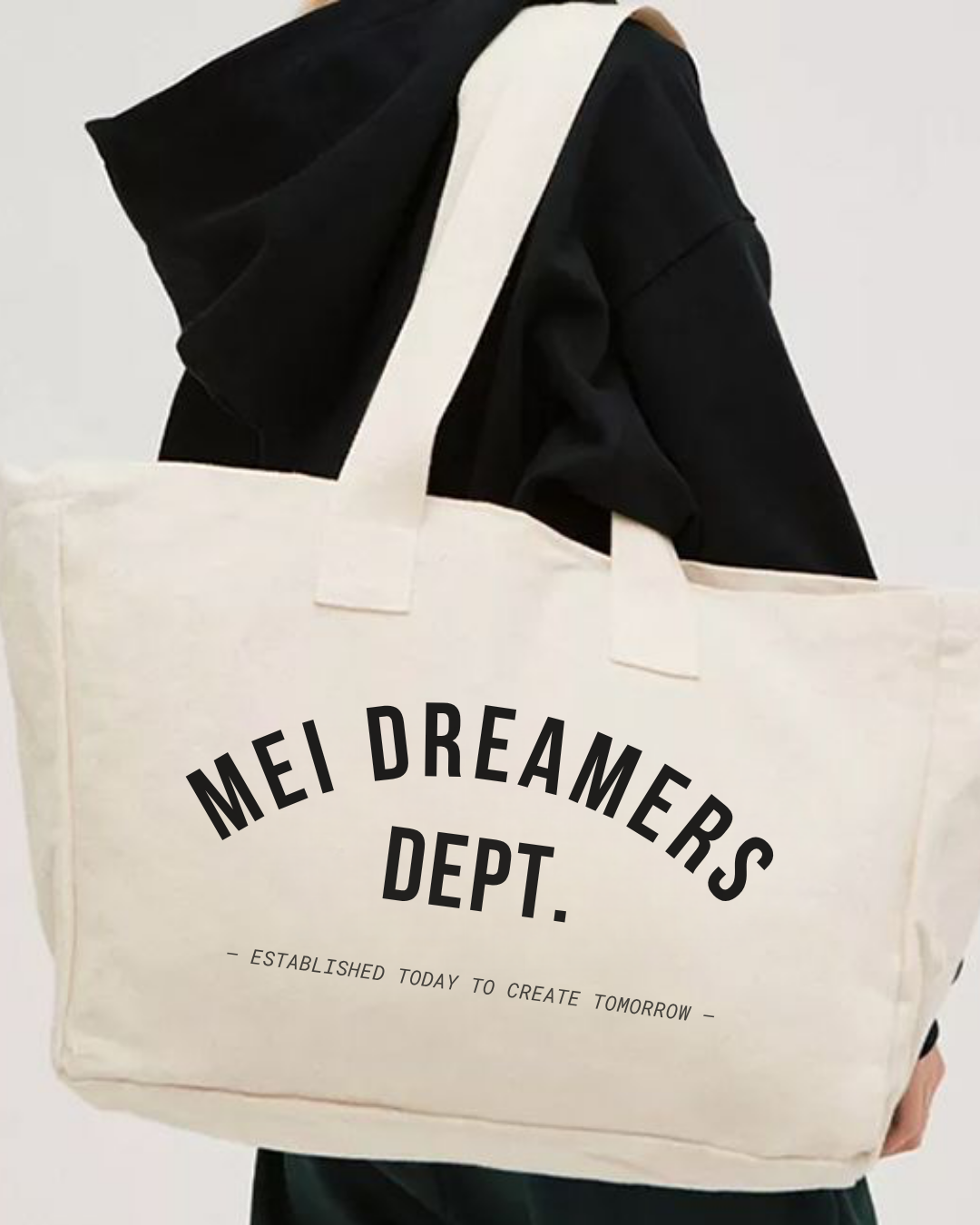 Person holding a cream-colored tote bag with black text that says "MEI DREAMERS DEPT." and smaller text beneath that reads "ESTABLISHED TODAY TO CREATE TOMORROW". The person is wearing a black hoodie.