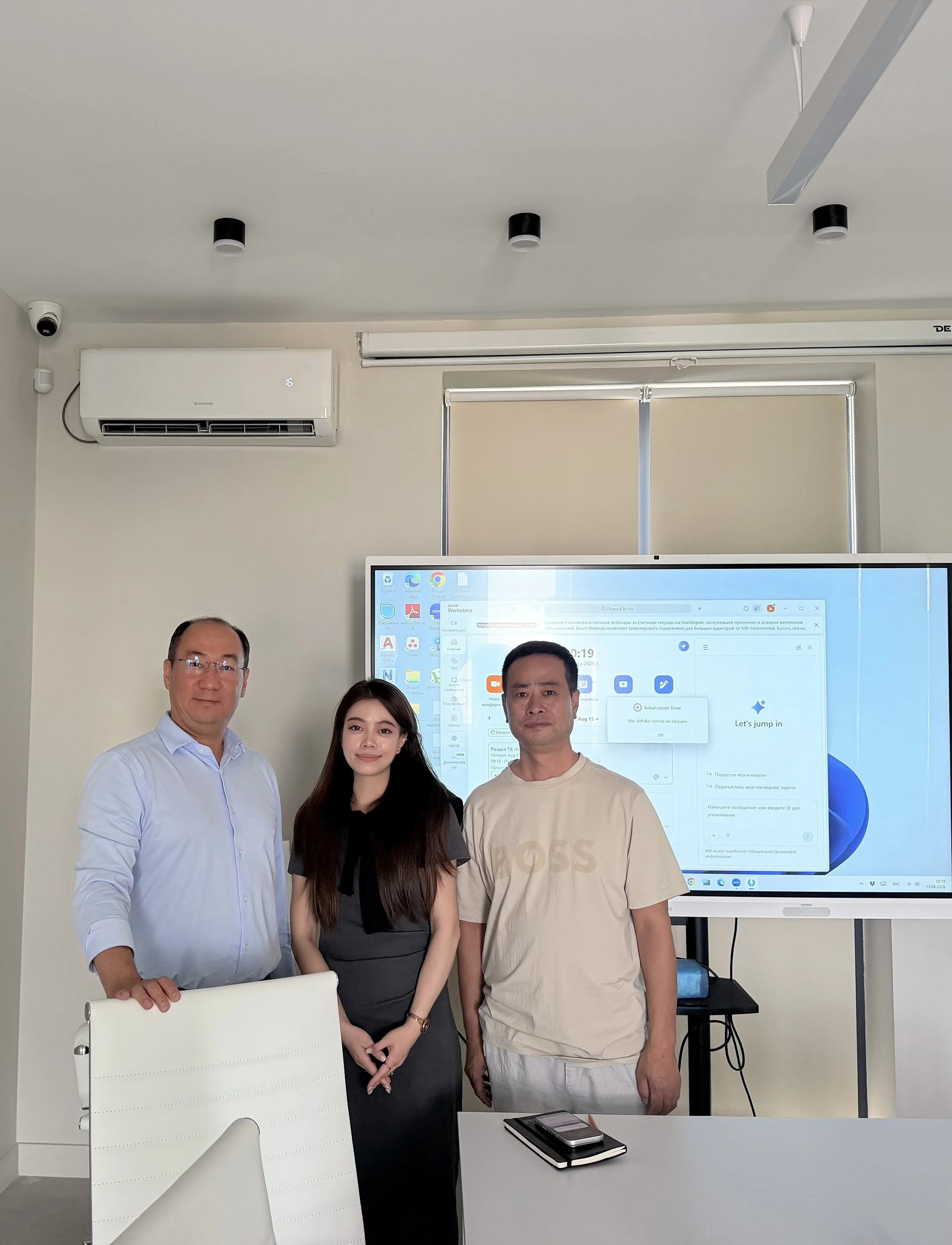 Four people standing in an office with a large screen in the background. The screen displays a computer desktop with various icons and open windows. The office has a white desk with a notebook, pen, and smartphone on it. The ceiling has a white air c